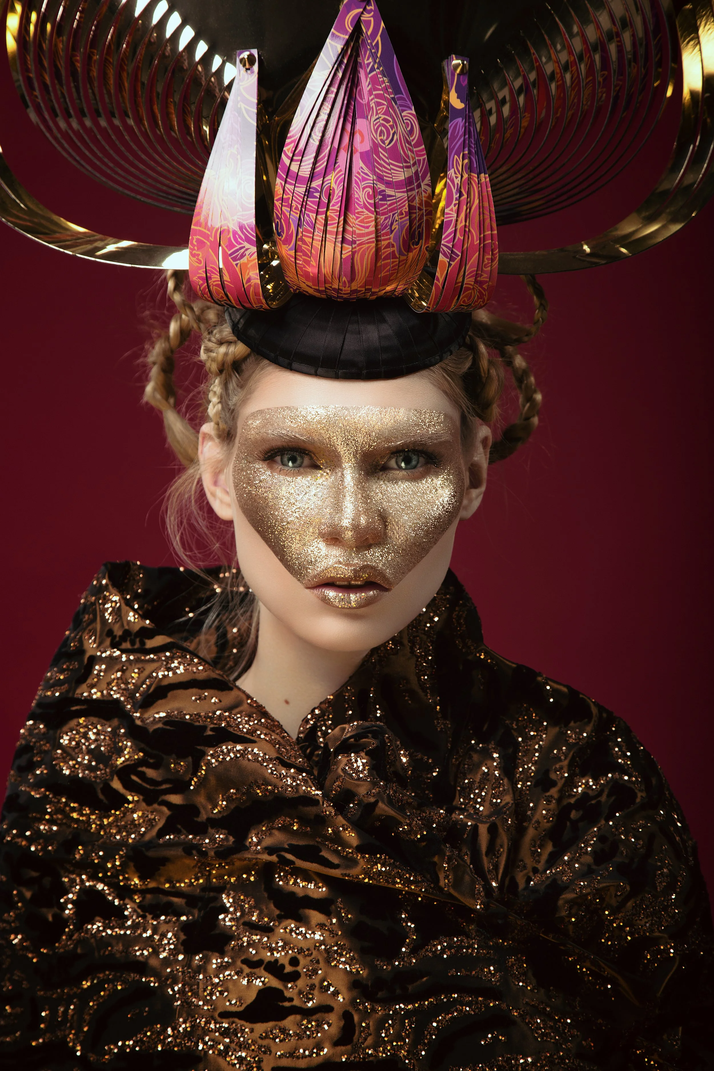 A woman with gold glitter on her face, wearing a black top and a gold and black patterned jacket. She has braided hair and is wearing an elaborate headpiece with a central purple and gold floral design, large black and gold circular structures, and g