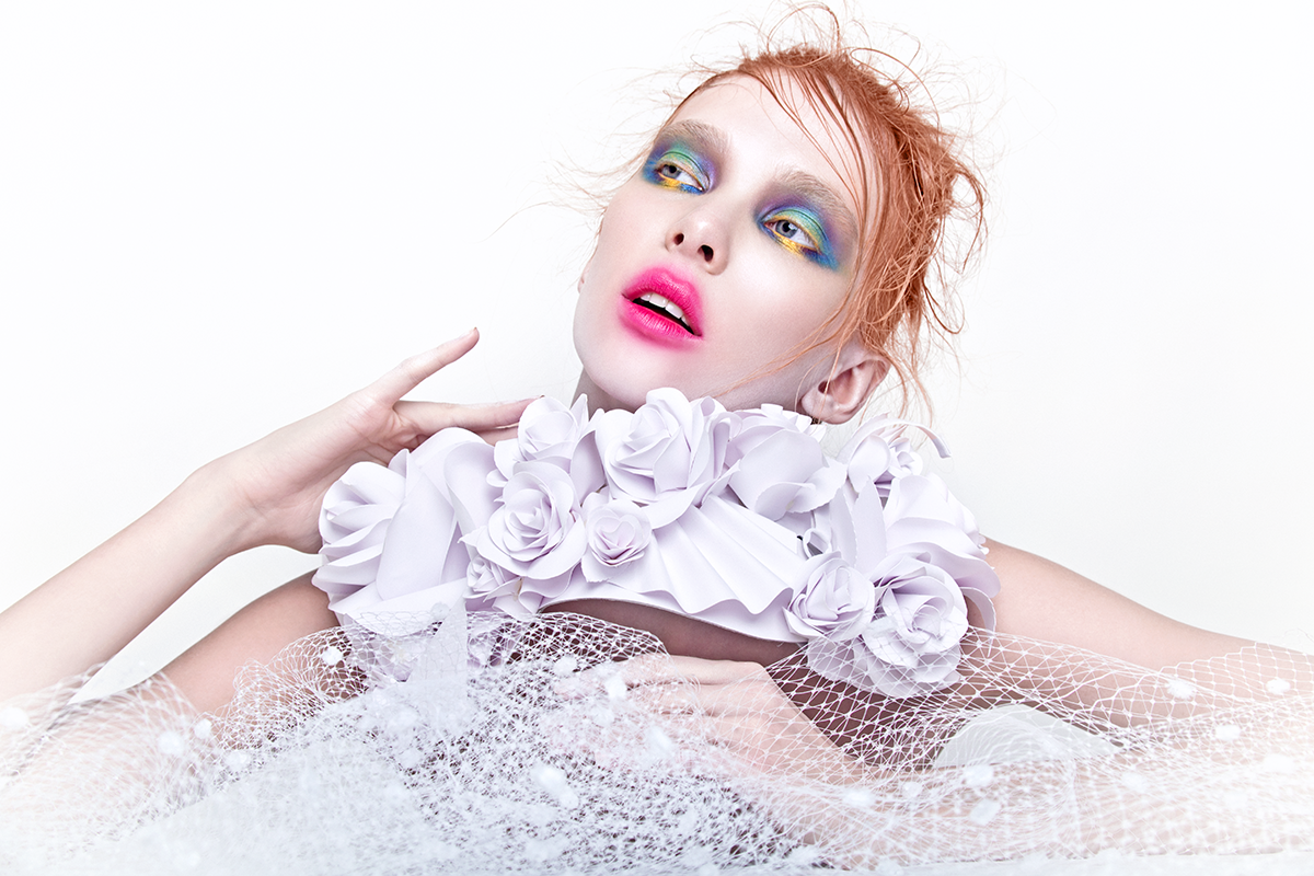 A woman with vibrant makeup, pink lips, and messy red hair wearing a white floral and netted fashion piece posing against a white background.