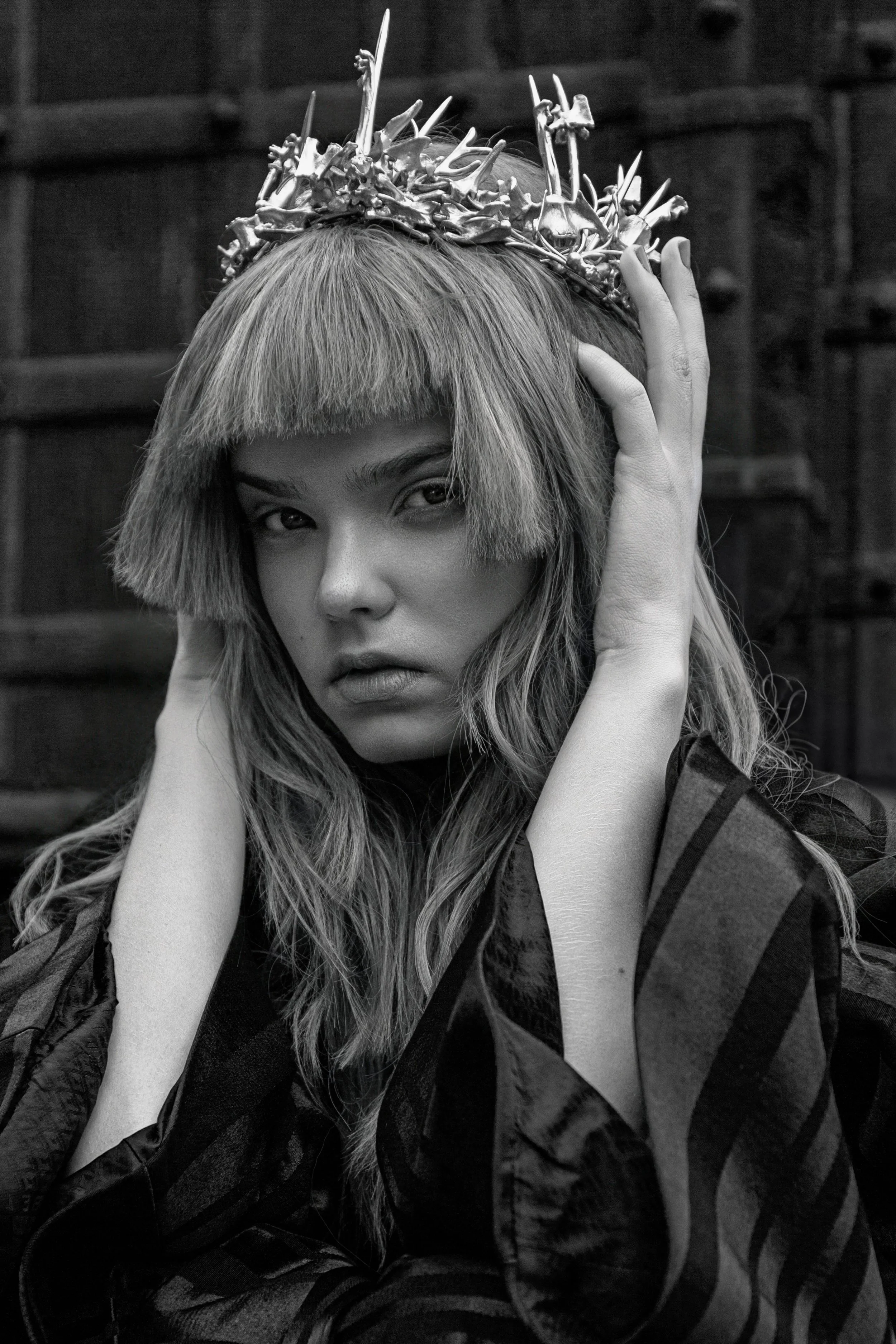 A black and white portrait of a woman wearing a crown made of swords and bones, with long hair, touching her face and looking at the camera.
