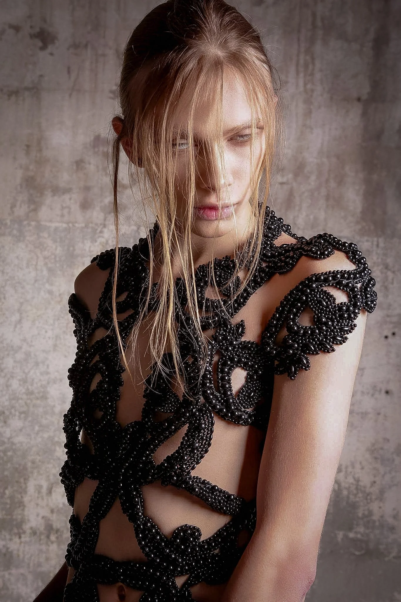 A woman with long blonde hair, styled with some strands hanging over her face, wearing intricate black beaded accessories on her arms and shoulders, posing against a textured gray wall.