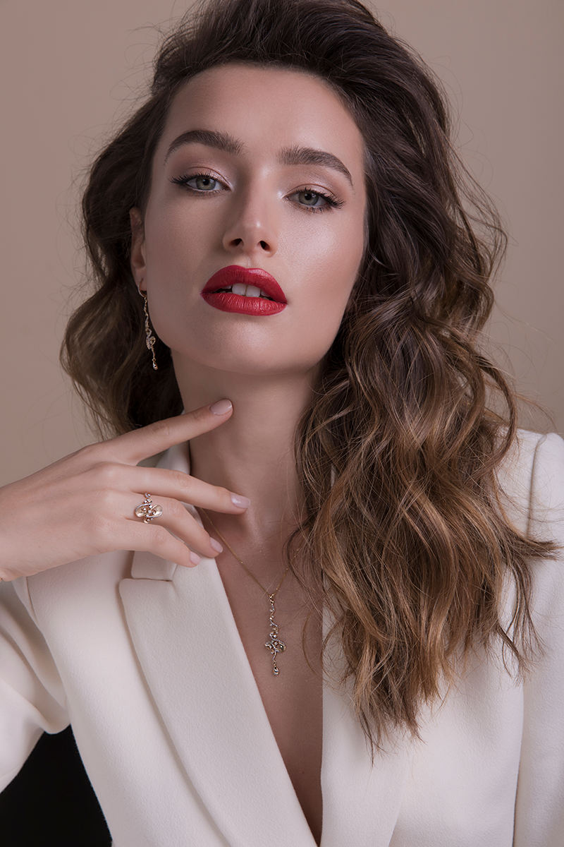 A young woman with wavy brown hair, wearing red lipstick, a white blazer, and jewelry, posing indoors with a neutral background.