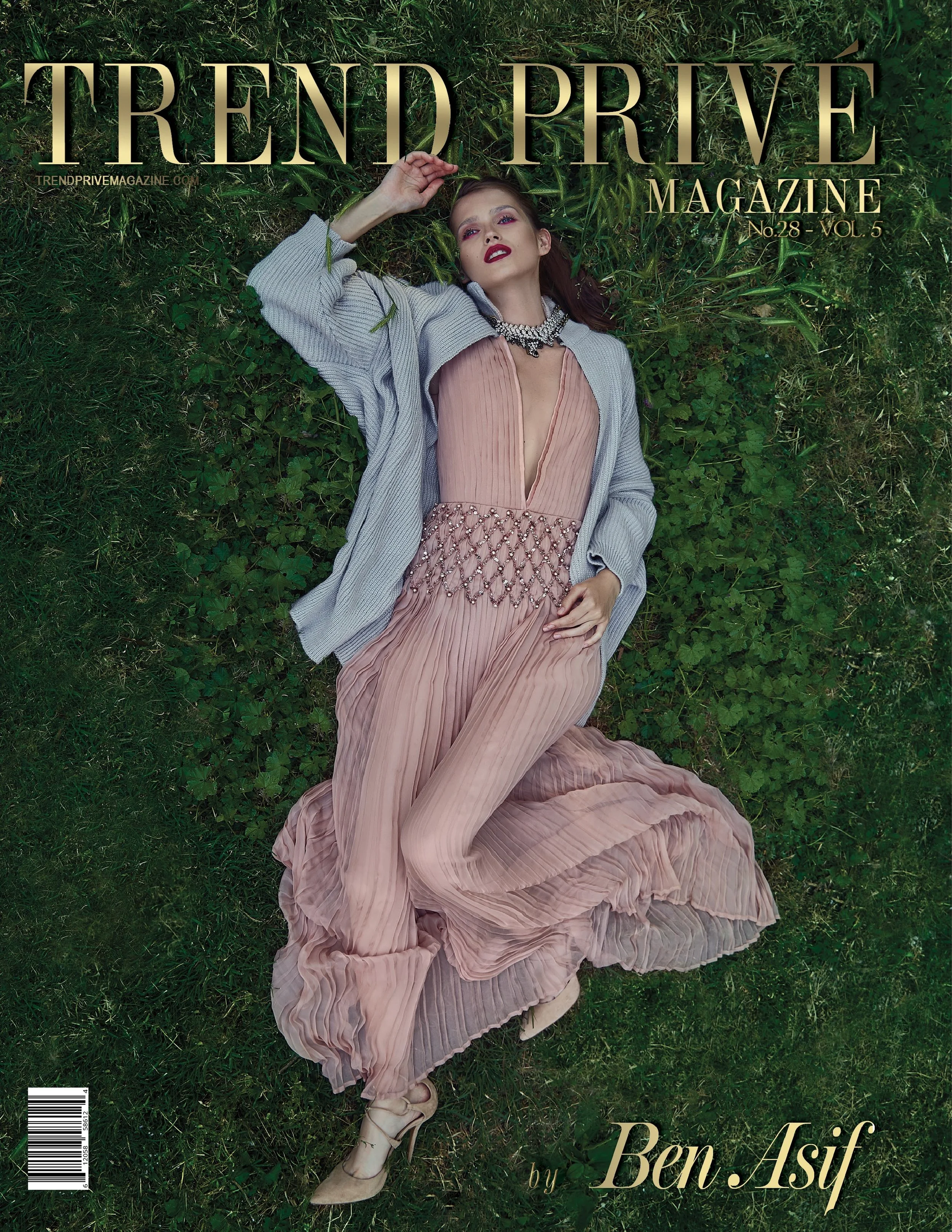 A woman in a pink pleated dress and gray cardigan lying on green grass, with red lipstick and jewelry, looking at the camera.
