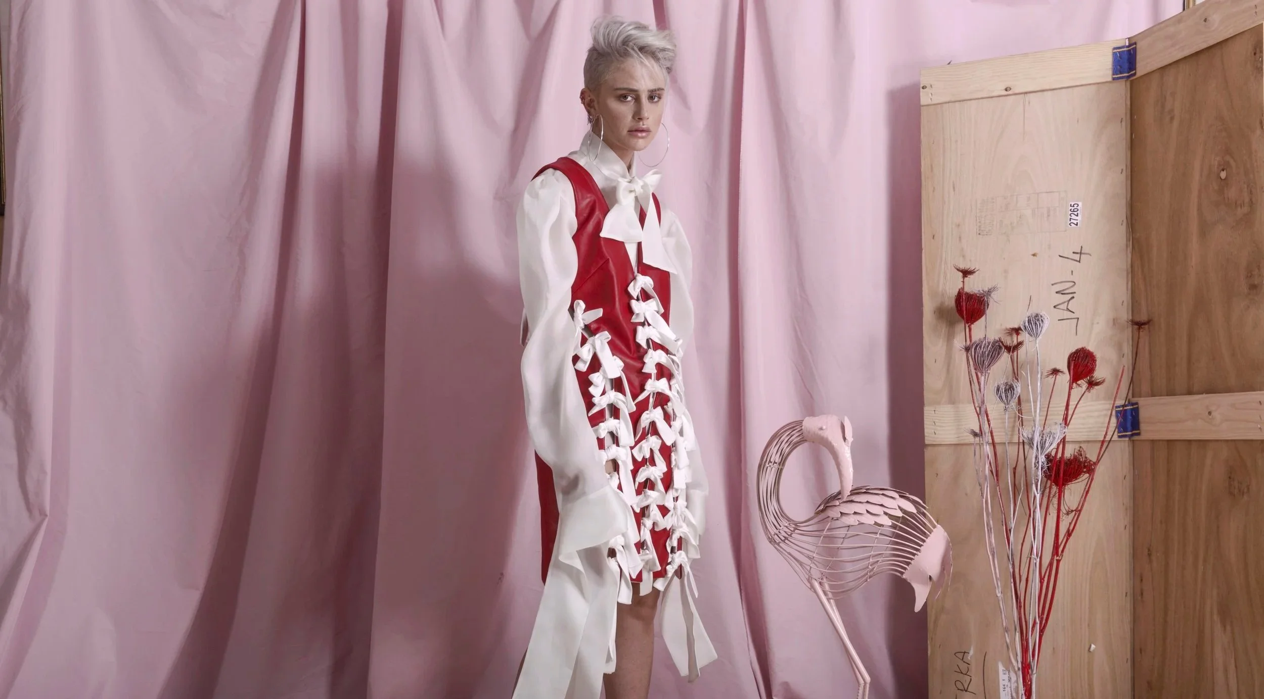 A woman with short platinum blonde hair and large hoop earrings posing in front of pink fabric backdrop, wearing a white blouse with a large bow and a red garment with white ribbon details, surrounded by pink and red decorative flowers and a wooden panel marked "JAN 4".