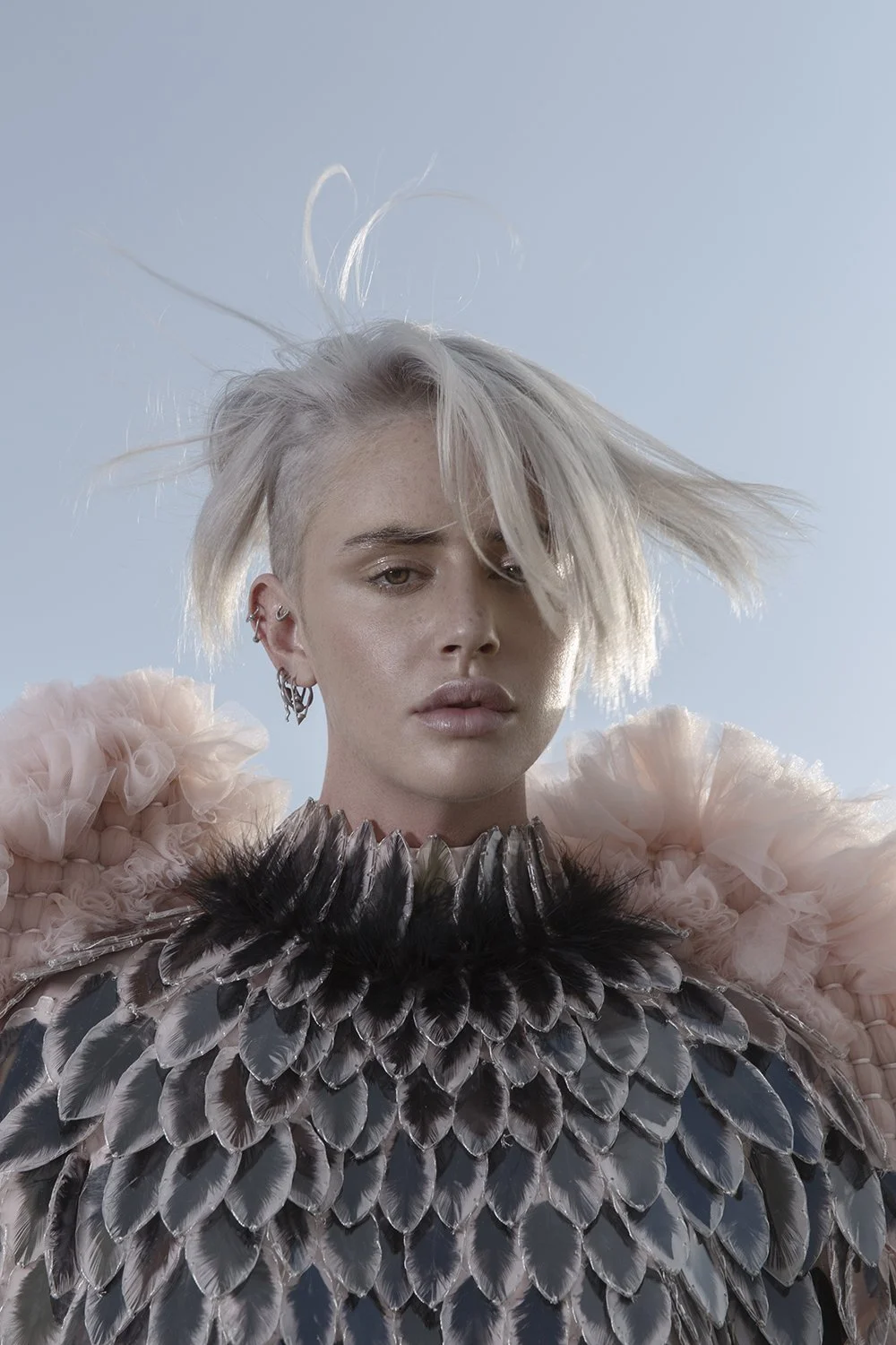A woman with platinum blonde hair, wearing a feathered outfit with black, silver, and pink feathers, and multiple earrings.