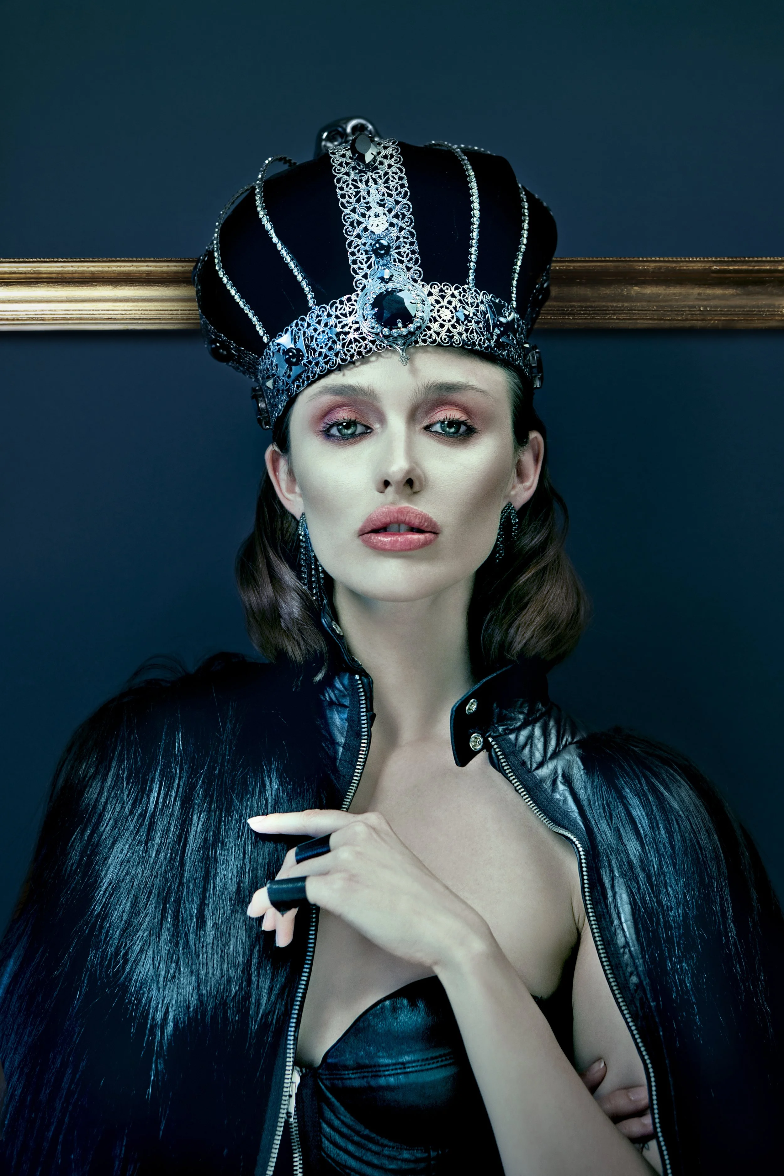 A woman wearing a large decorative crown with black and silver details, a black leather jacket, and a strapless black top. She has long, glossy black hair and light makeup, with a dark wall and a gold horizontal accent in the background.