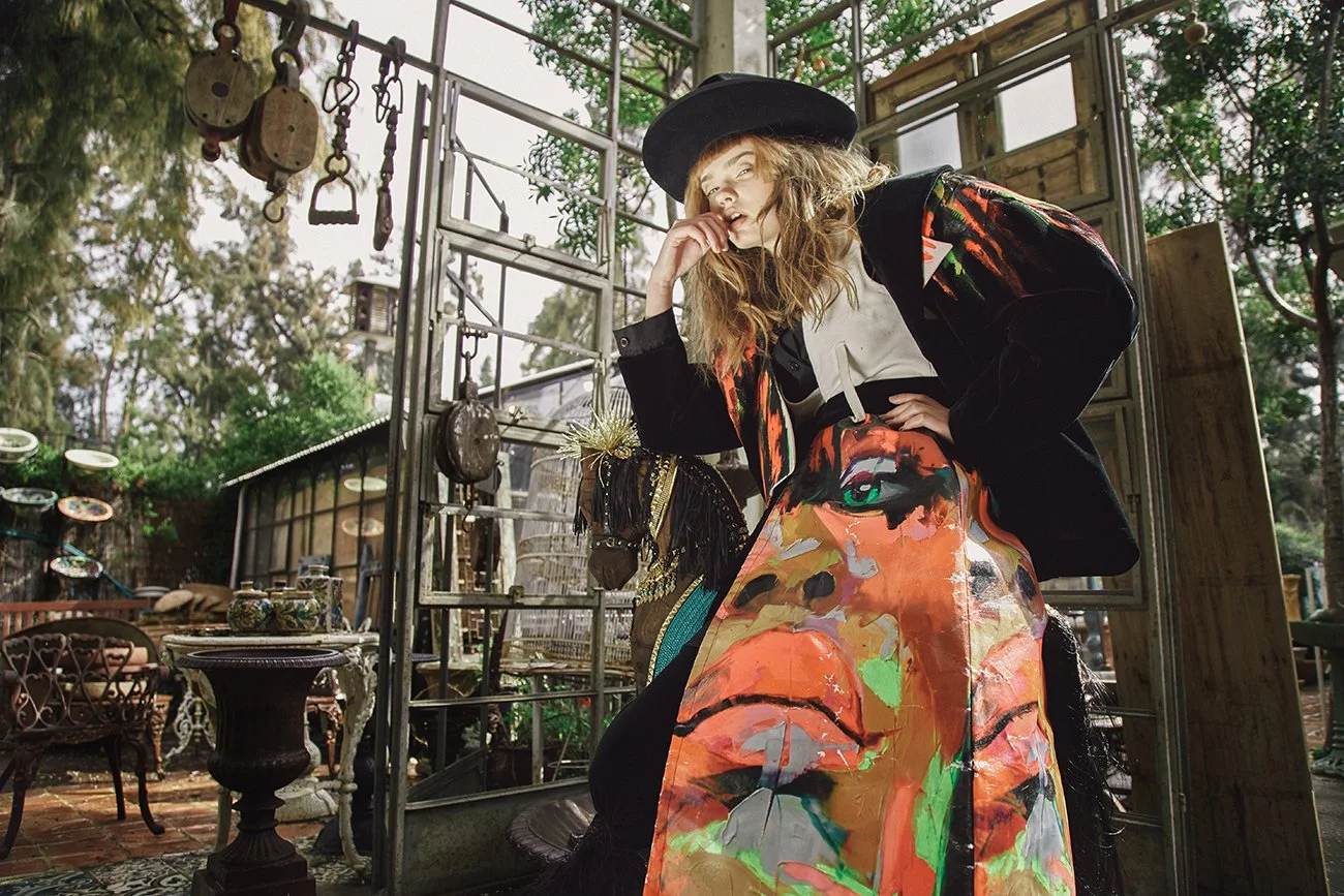 A woman with wavy hair wearing a black wide-brimmed hat, a black jacket with colorful patterns, white top, and a long skirt with a vibrant face print, is posing with a thoughtful expression in an outdoor setting filled with eclectic furniture and dec