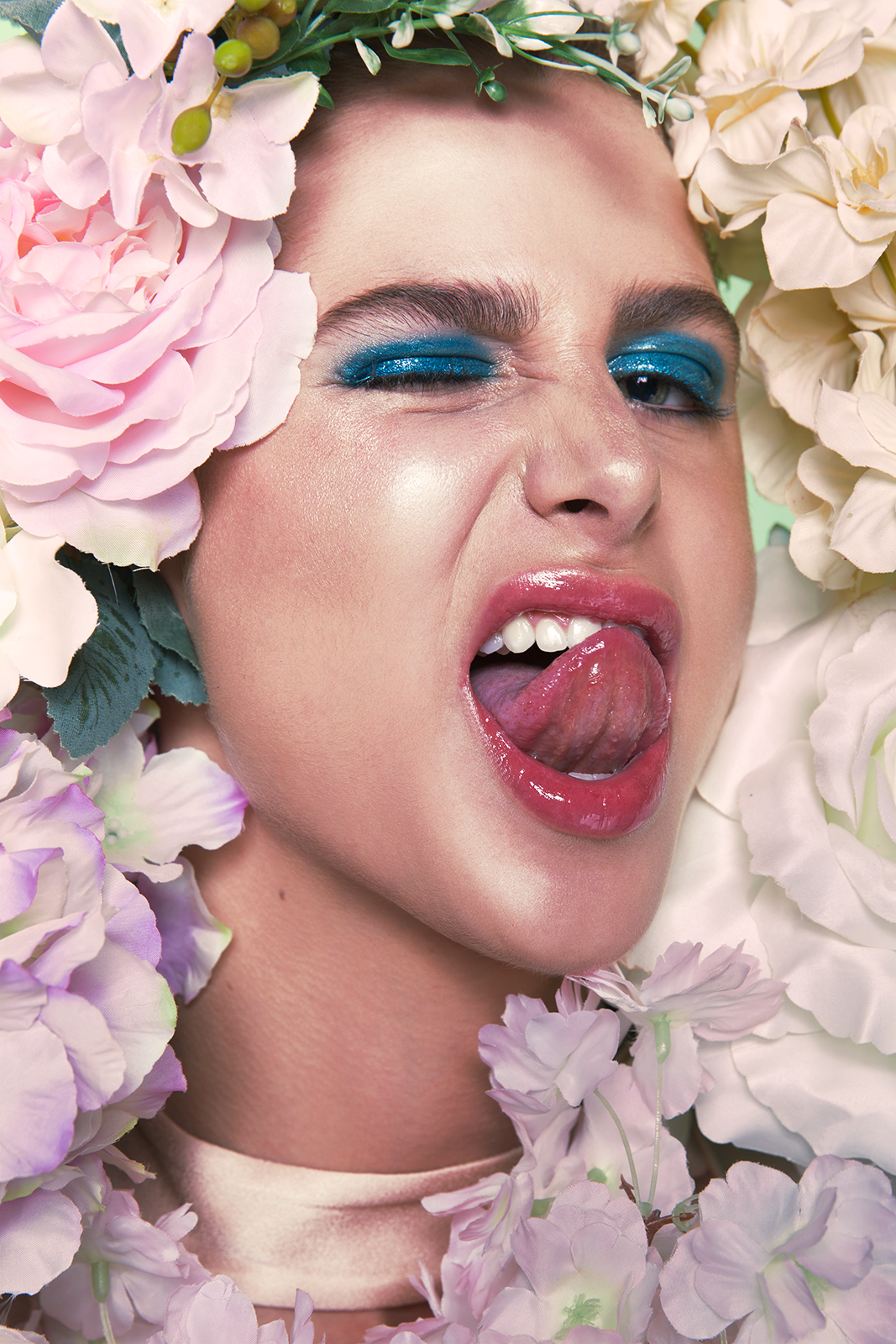 close-up of a woman's face with floral crown, blue eye makeup, and shiny lipstick, making a playful expression with her tongue out.