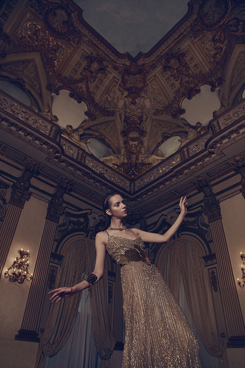 A woman in a luxurious, gold, sequined evening gown standing in an opulent room with ornate ceiling decorations, chandeliers, and draped curtains.