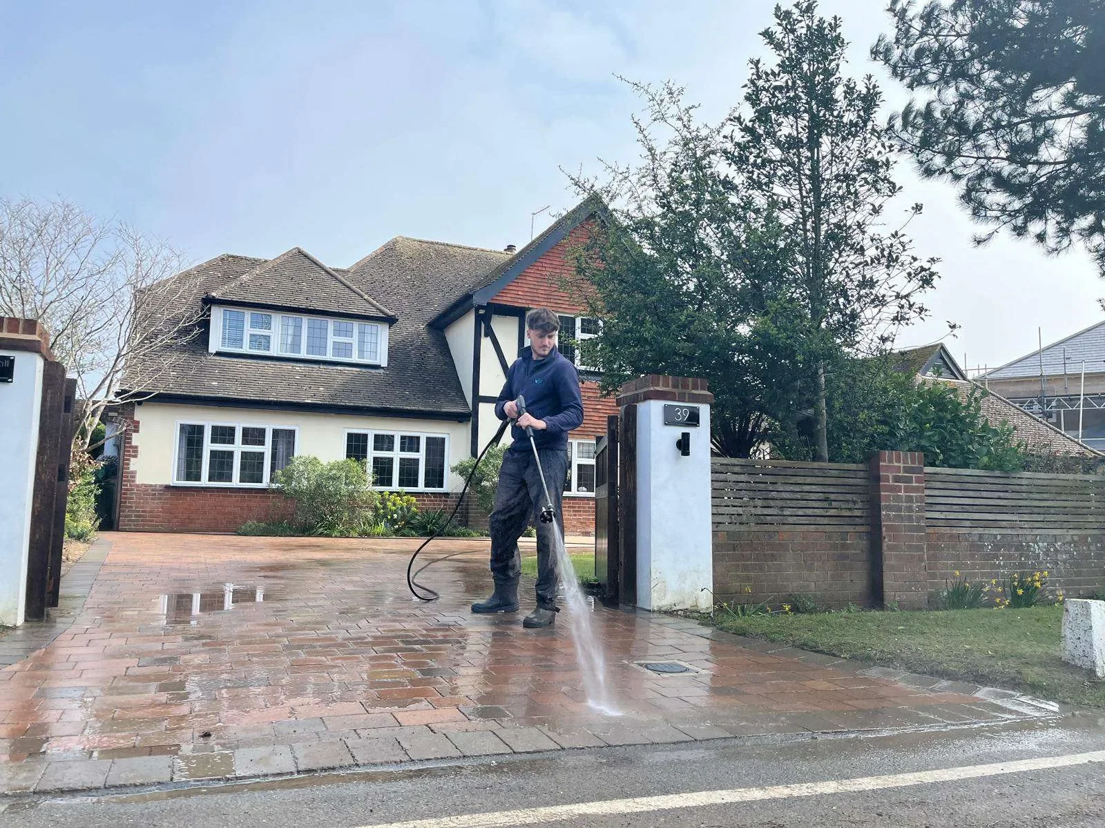 A person pressure washing a brick driveway in front of a house with a white and black exterior, surrounded by trees and a brick fence.