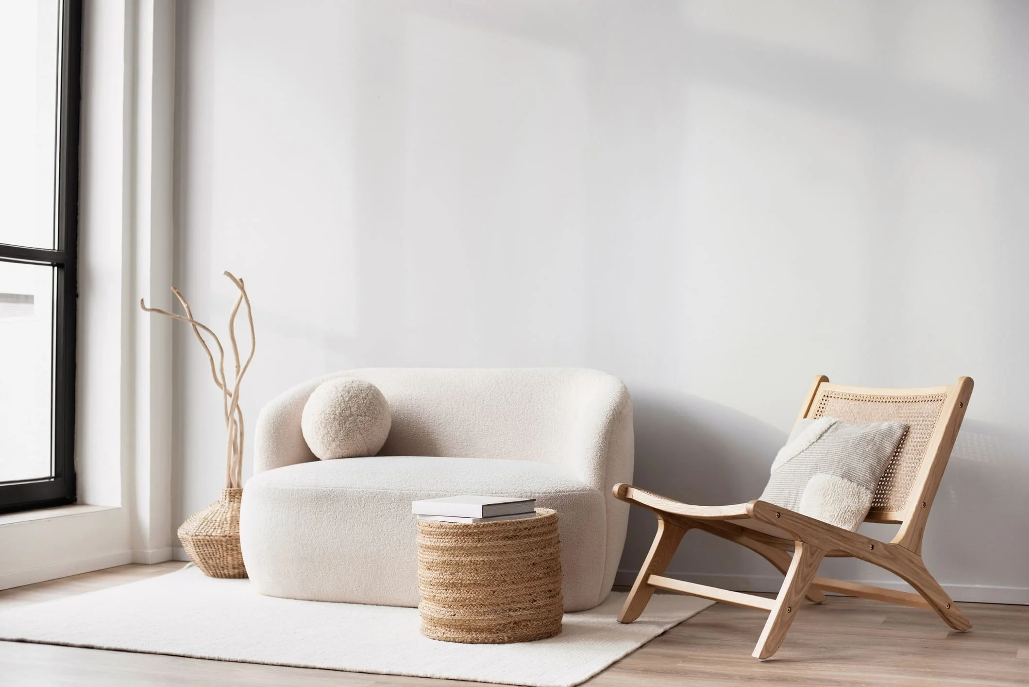 Salon minimaliste avec canapé blanc, fauteuil en bois, coussins, et décor en fibres naturelles, près de grande fenêtre avec rideaux blancs.