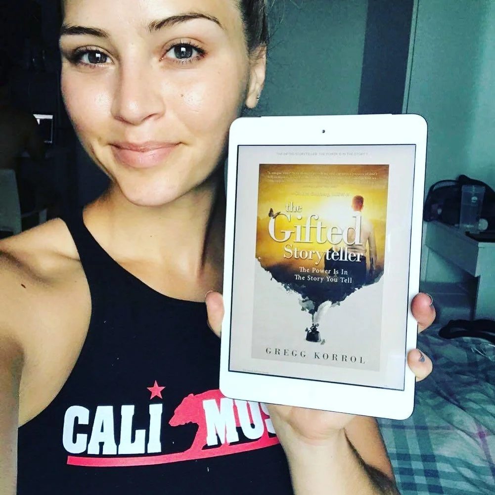 A woman with short brown hair, wearing a black tank top with red and white text, smiling and holding a white tablet displaying the cover of the book 'The Gifted Storyteller' by Gregg Korrol. The background shows a room with a bed and some furniture.