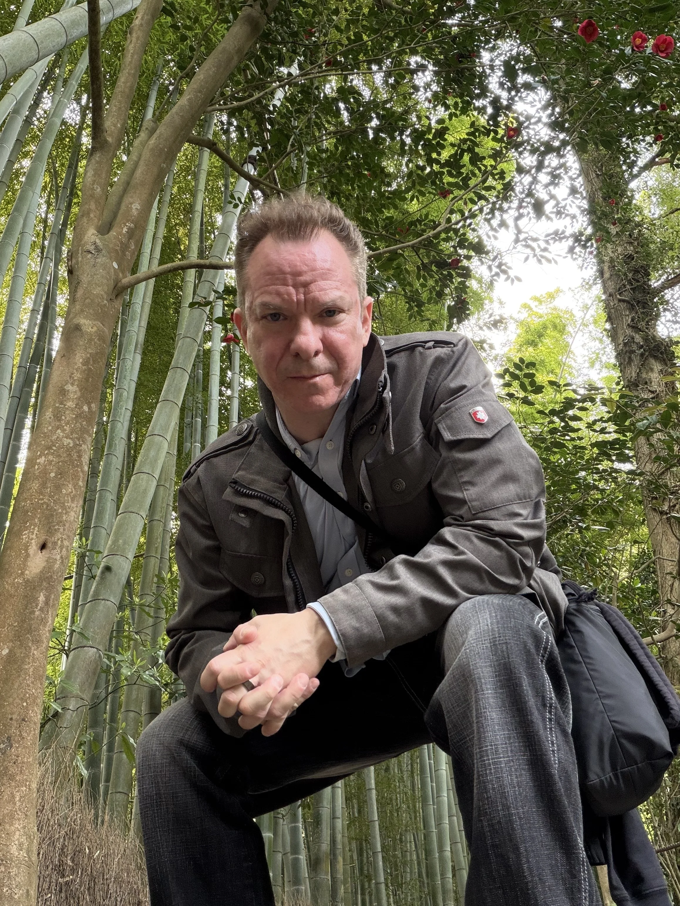A man crouching in a bamboo forest, wearing a gray jacket and gray trousers, with foliage and bamboo stalks behind him. Gregg Korrol- The Gifted Storyteller. Create the life you've been waiting for and dreaming of.