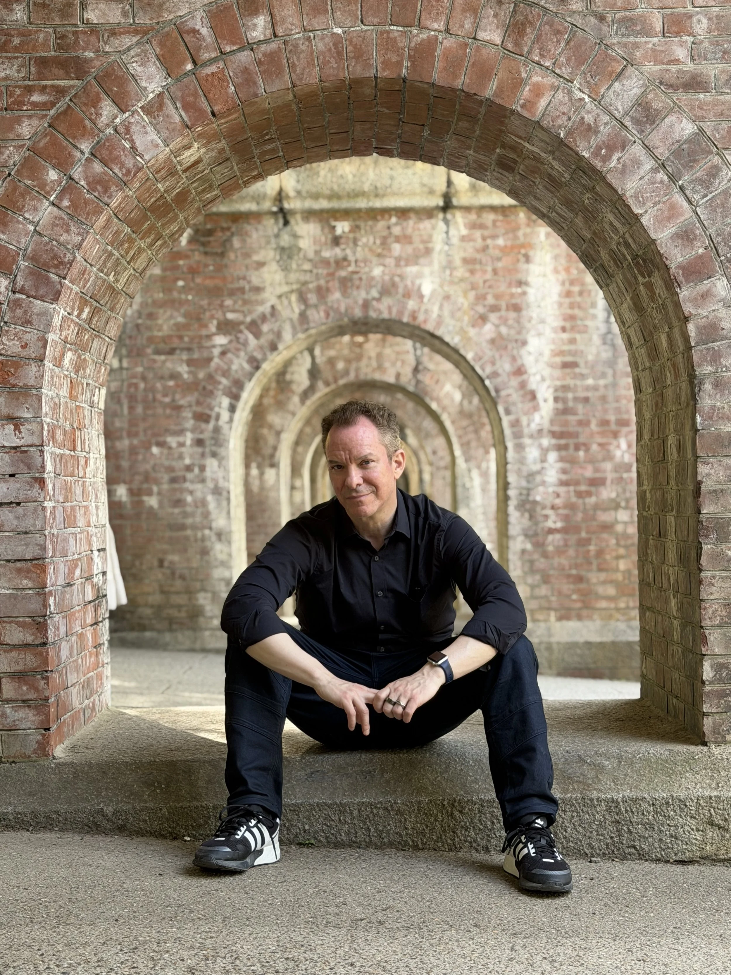 A man crouching under an archway made of red bricks, with a series of arches extending into the background. Gregg Korrol- The Gifted Storyteller. Create the life you've been waiting for and dreaming of.