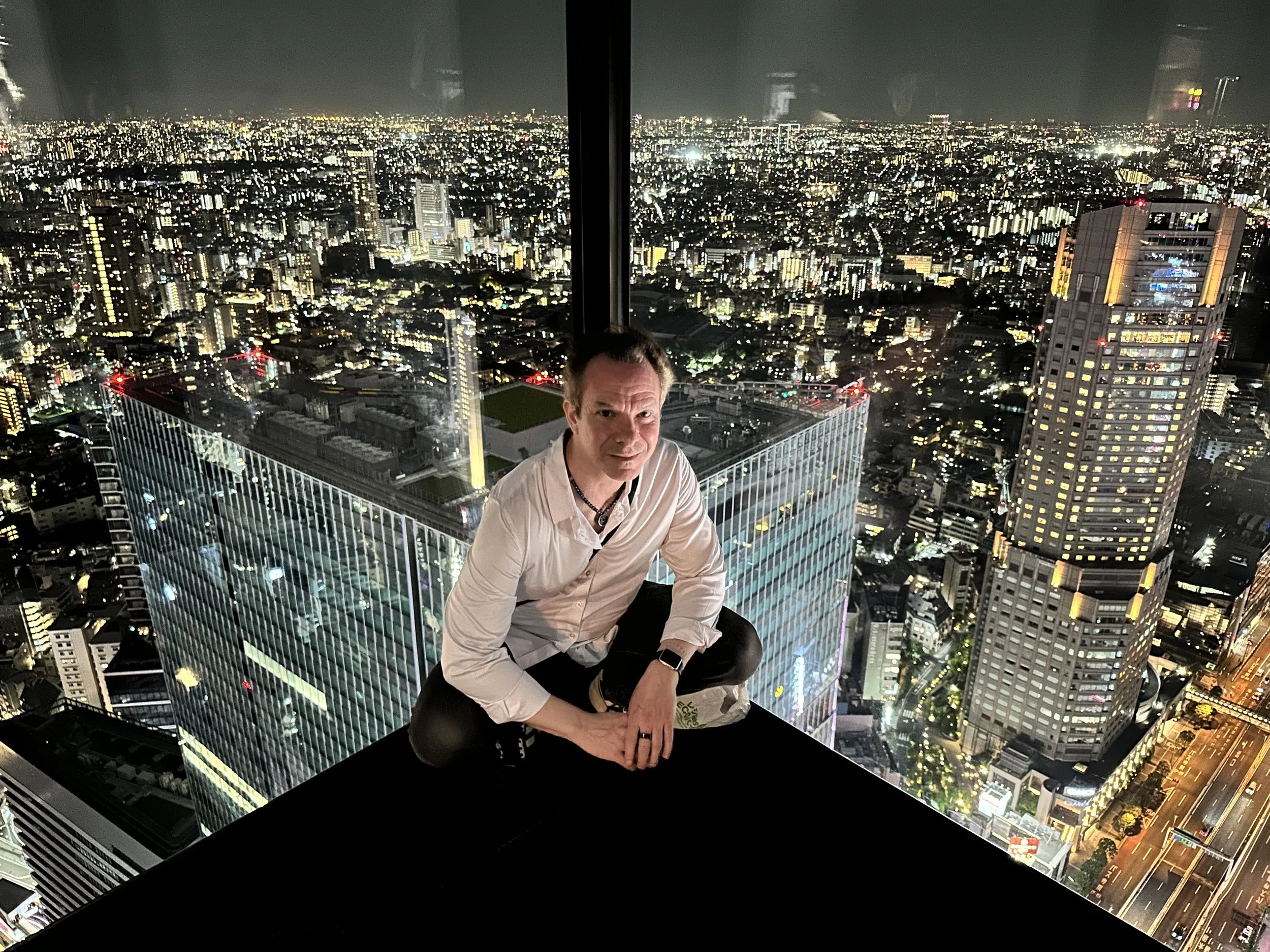 A man in a white shirt and black pants, kneeling on a glass ledge of a skyscraper at night with a city skyline illuminated in the background. Gregg Korrol- The Gifted Storyteller. Create the life you've been waiting for and dreaming of.