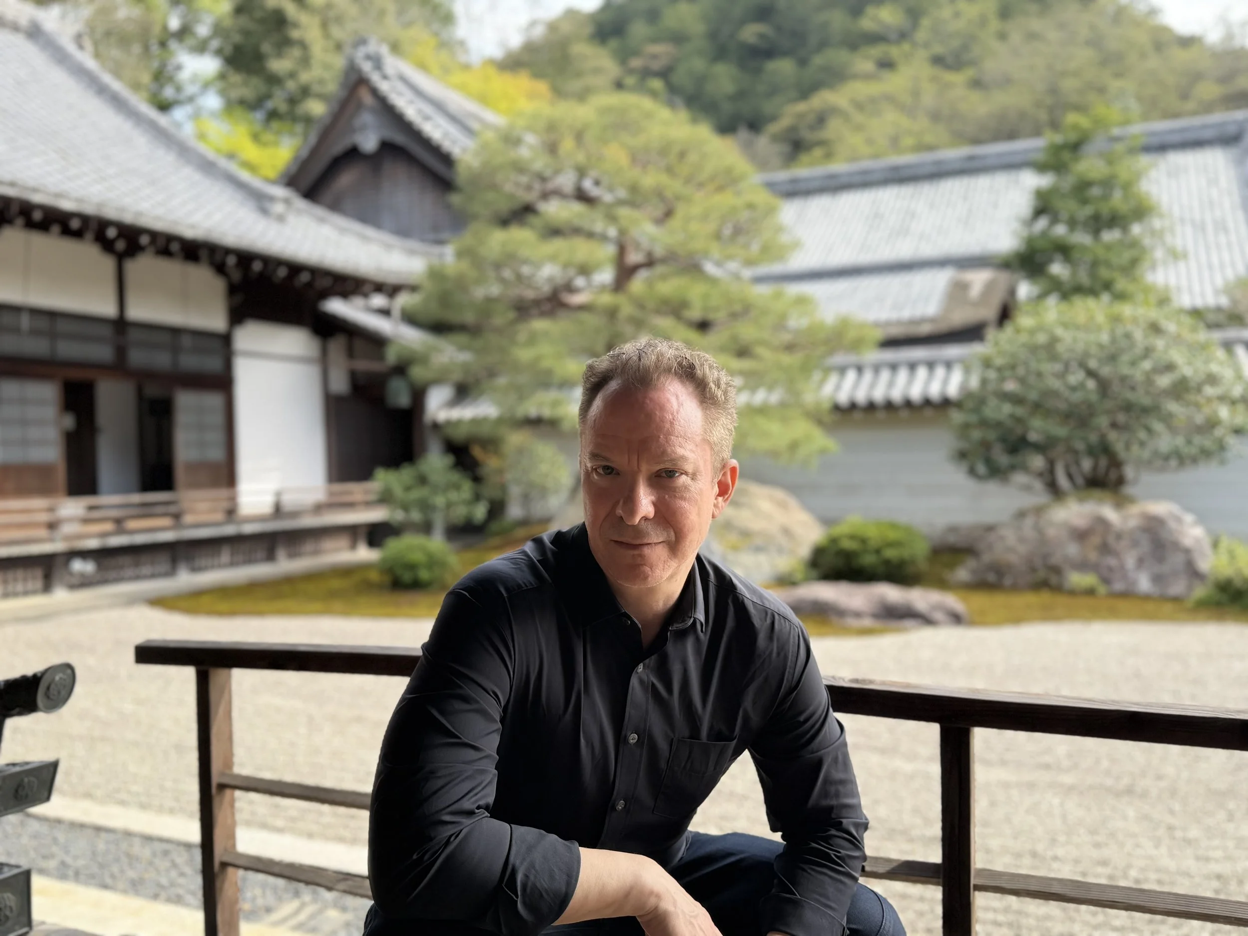 A man with short hair and a black shirt in a traditional Japanese garden with pagoda-style buildings, trees, and rocks in the background. Gregg Korrol- The Gifted Storyteller. Create the life you've been waiting for and dreaming of.