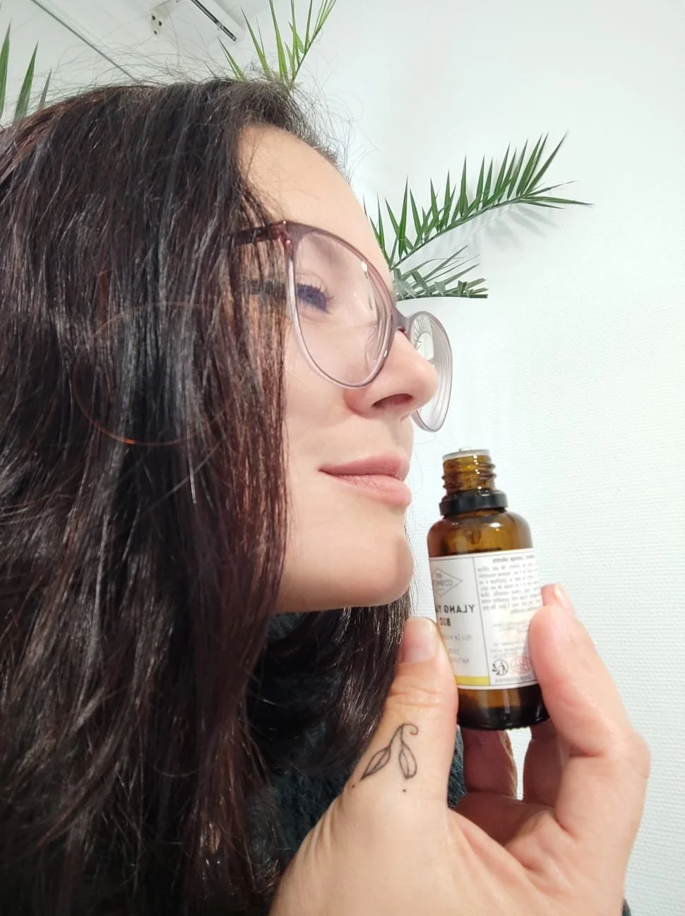 Donna con occhiali chiari e capelli lunghi, che tiene una bottiglia di integratori o medicine in mano, con uno sfondo di parete bianca e piante verdi.