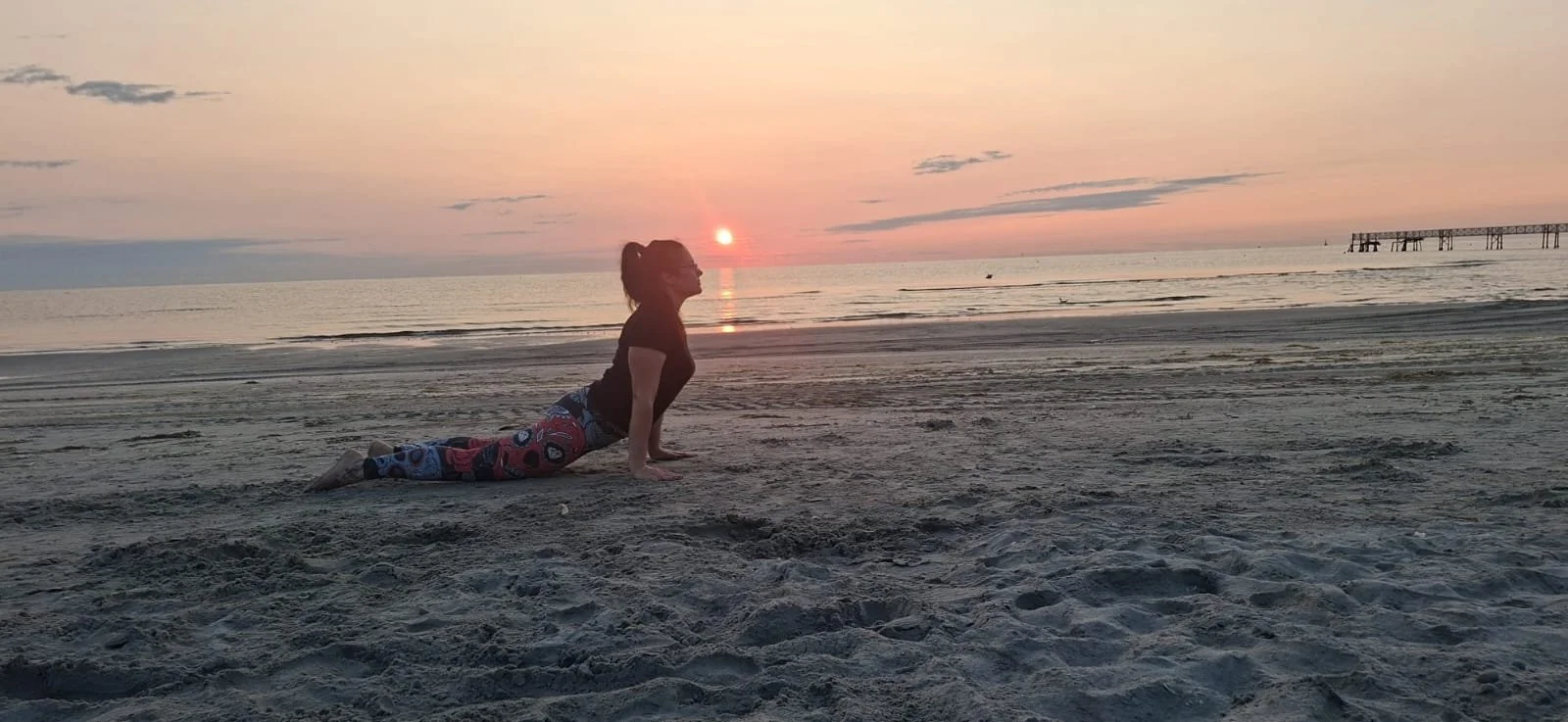 Donna fa esercizi di yoga o stretching sulla spiaggia al tramonto, con il mare e il pontile sullo sfondo.