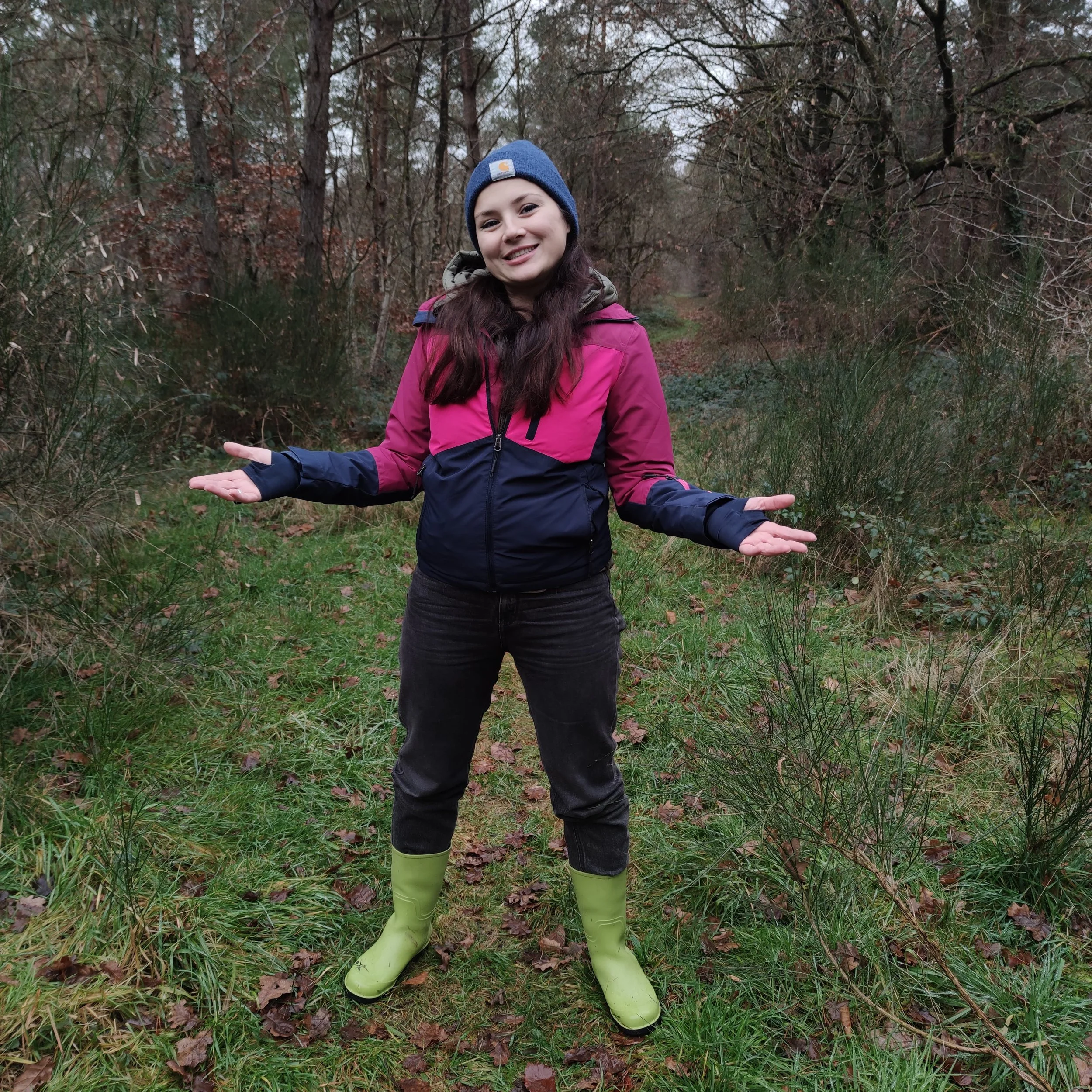 Una ragazza sorridente vestita con giacca rosa e pantaloni neri, indossa un berretto blu e stivali verdi in una foresta autunnale con foglie sul terreno.