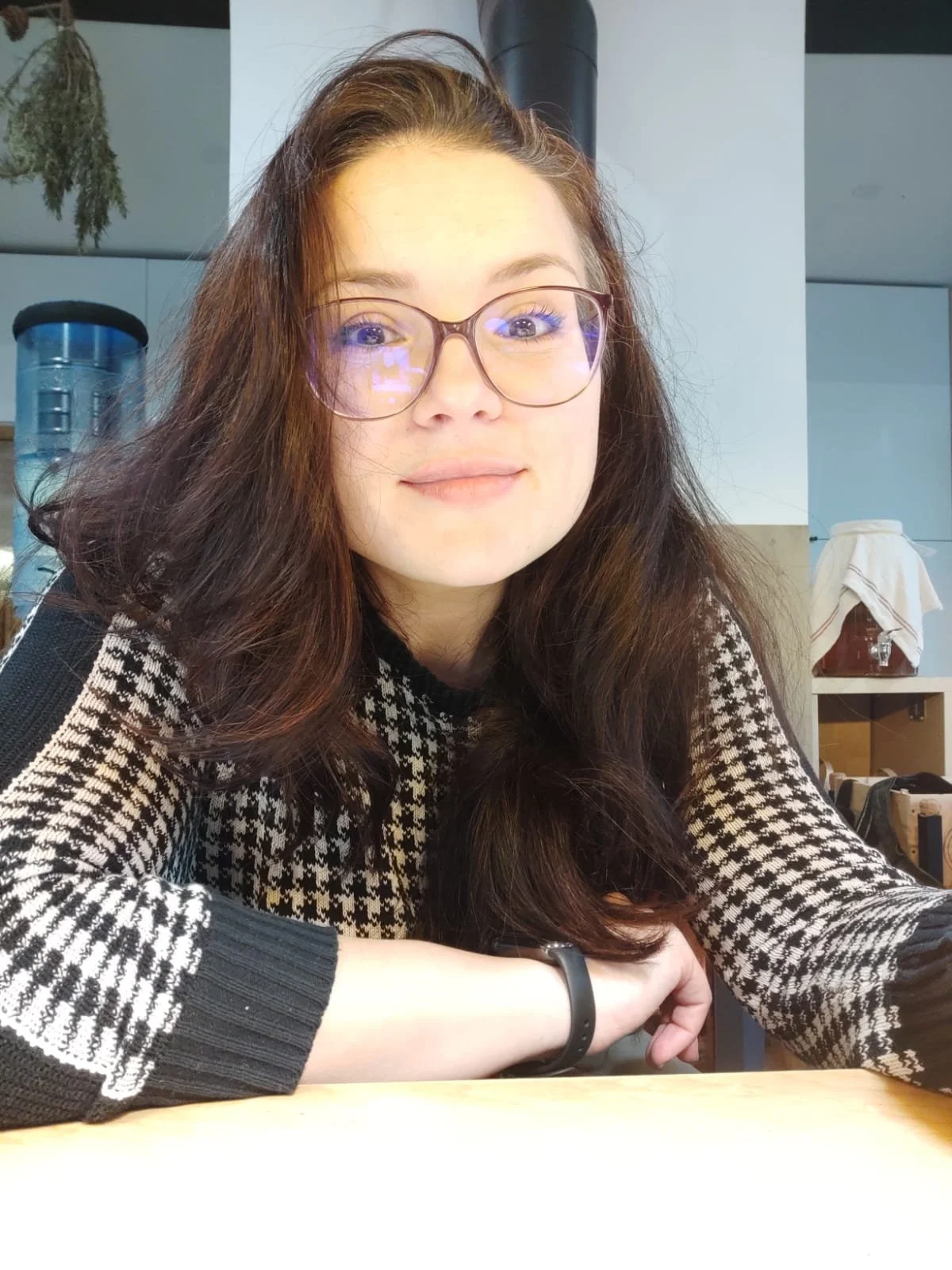 Una donna con lunghi capelli castani e occhiali rossi, seduta a un tavolo in un ambiente interno, che sorride lievemente.