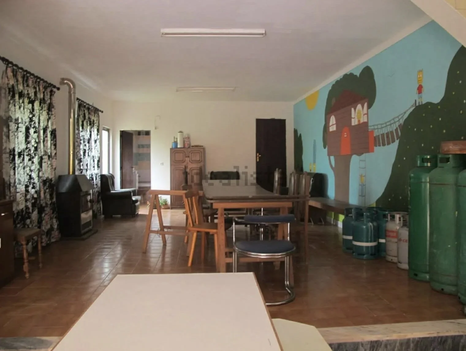 Interior of a room with wooden flooring, a colorful painted mural of a treehouse and landscape on the wall, multiple tables and chairs, a cabinet, window with floral curtains, and portable gas tanks on the right.
