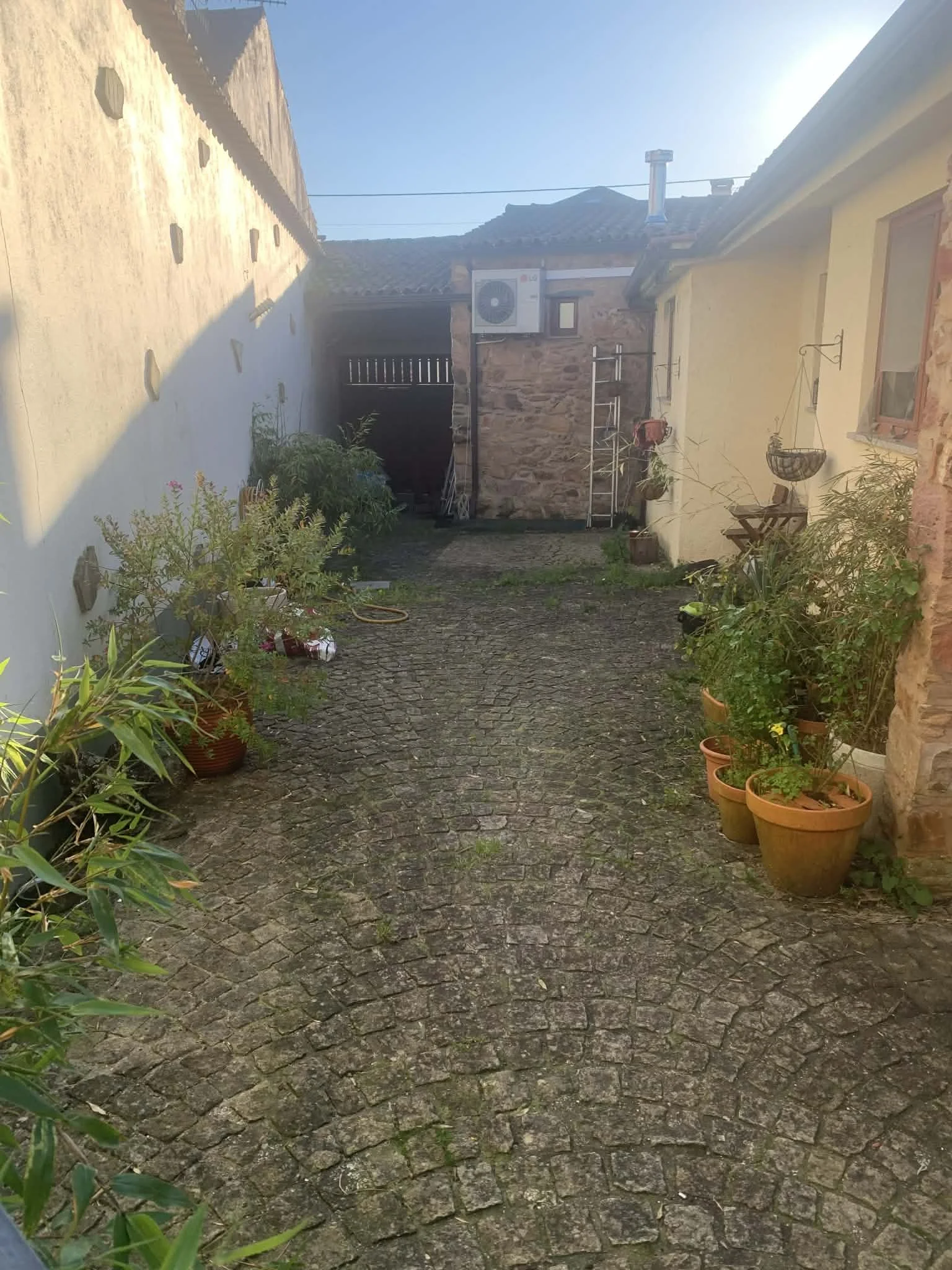 A narrow cobblestone alleyway with potted plants along the sides and a yellow house on the right. There are old walls and a small structure at the end, with outdoor items and herding equipment.
