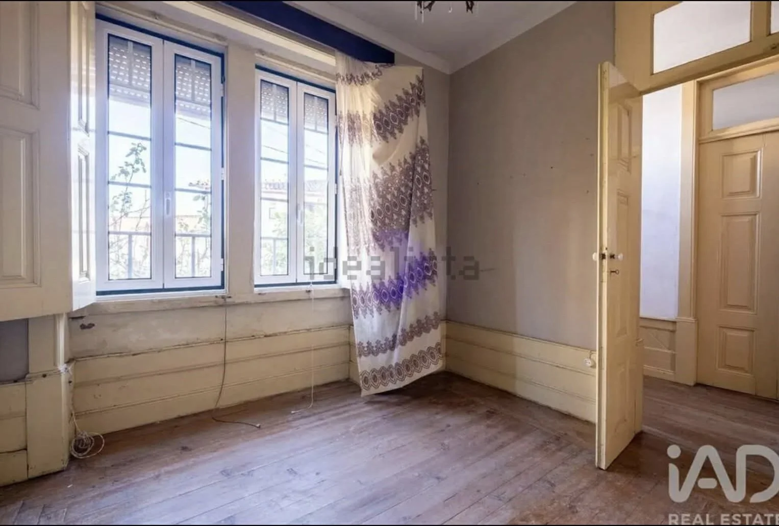 Empty room with wooden floors, large windows with white frames, a patterned curtain, and an open wooden door leading to another room.