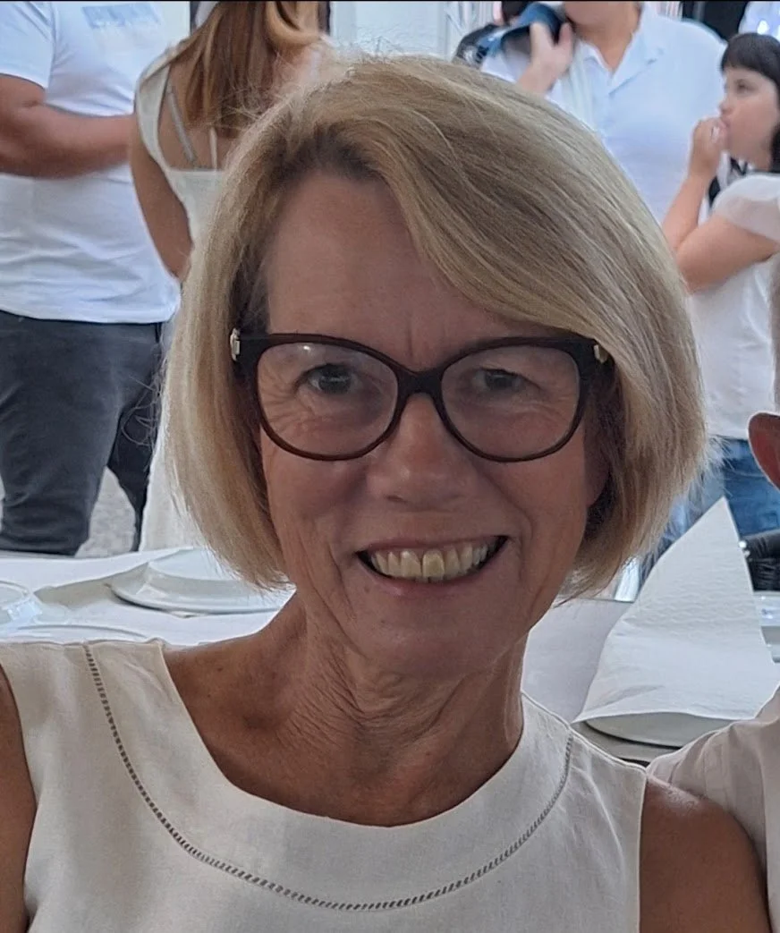 A smiling elderly woman with short blonde hair and glasses, sitting at a table with plates and napkins, at a social gathering with people in the background.