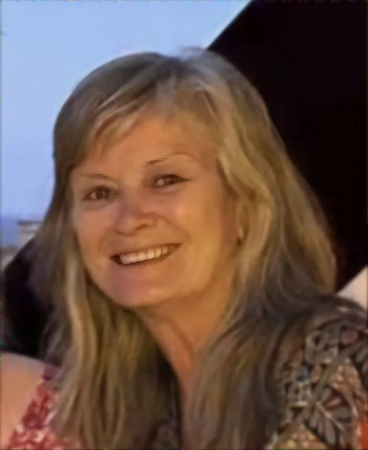A smiling woman with blonde hair and light skin, dressed in a patterned top, in a close-up portrait.