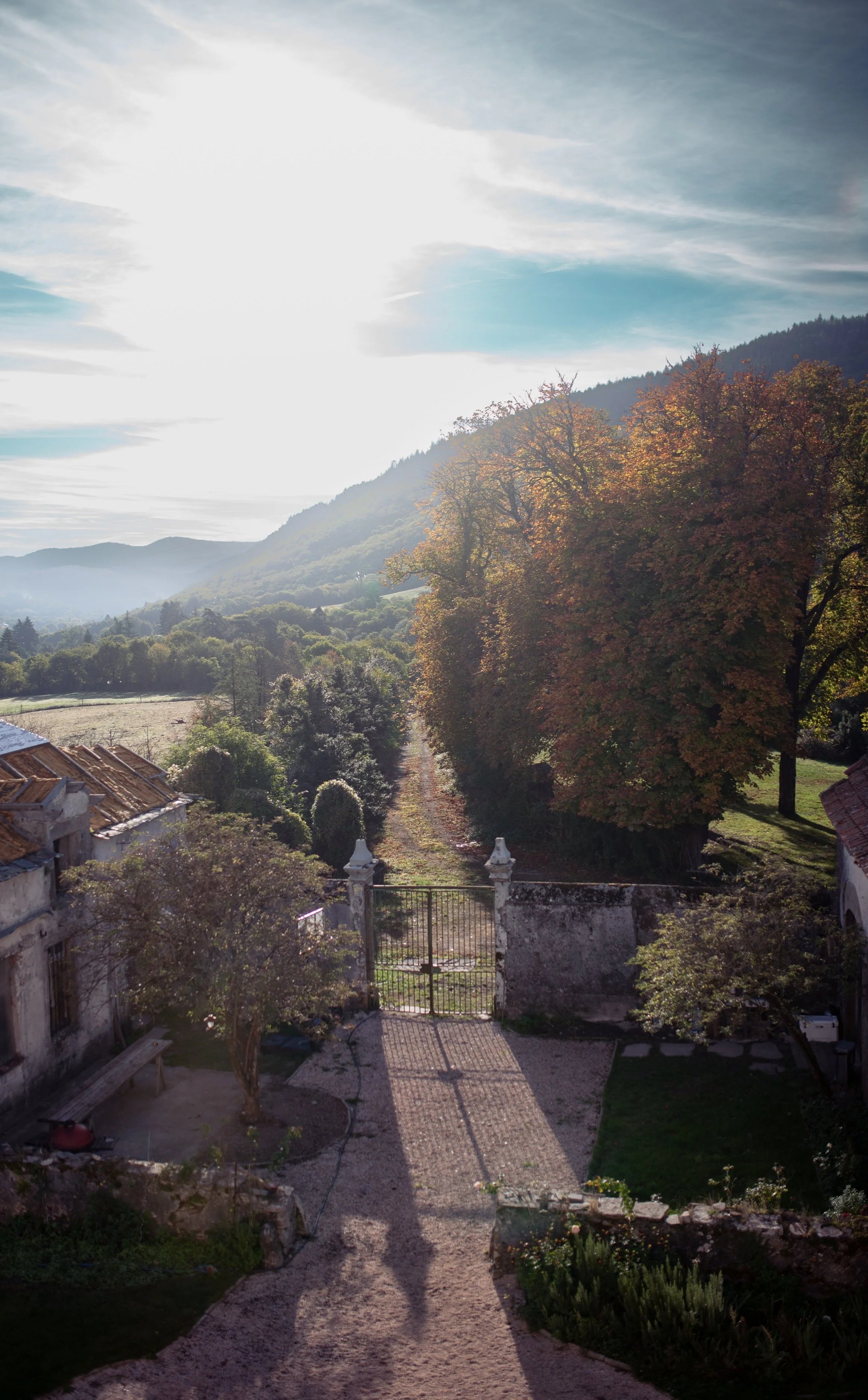 A rural scene with a dirt pathway leading to a gate, surrounded by trees with fall foliage, old buildings, and distant hills under a partly cloudy sky with bright sunlight.