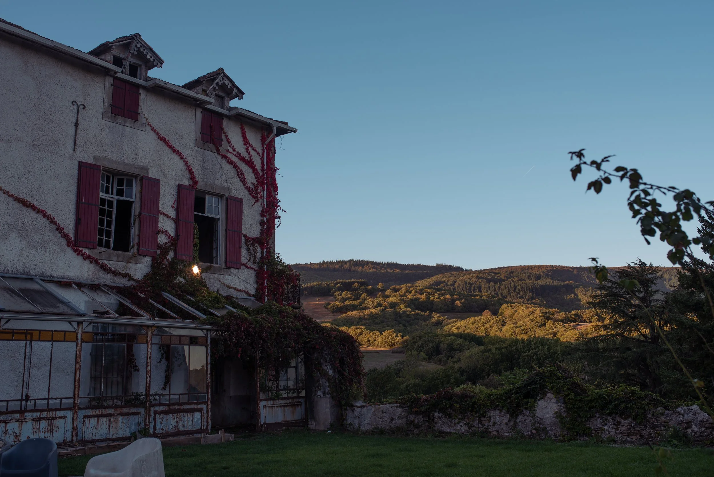 An old white house with red shutters and vines, set against a backdrop of rolling hills and a clear blue sky at sunset