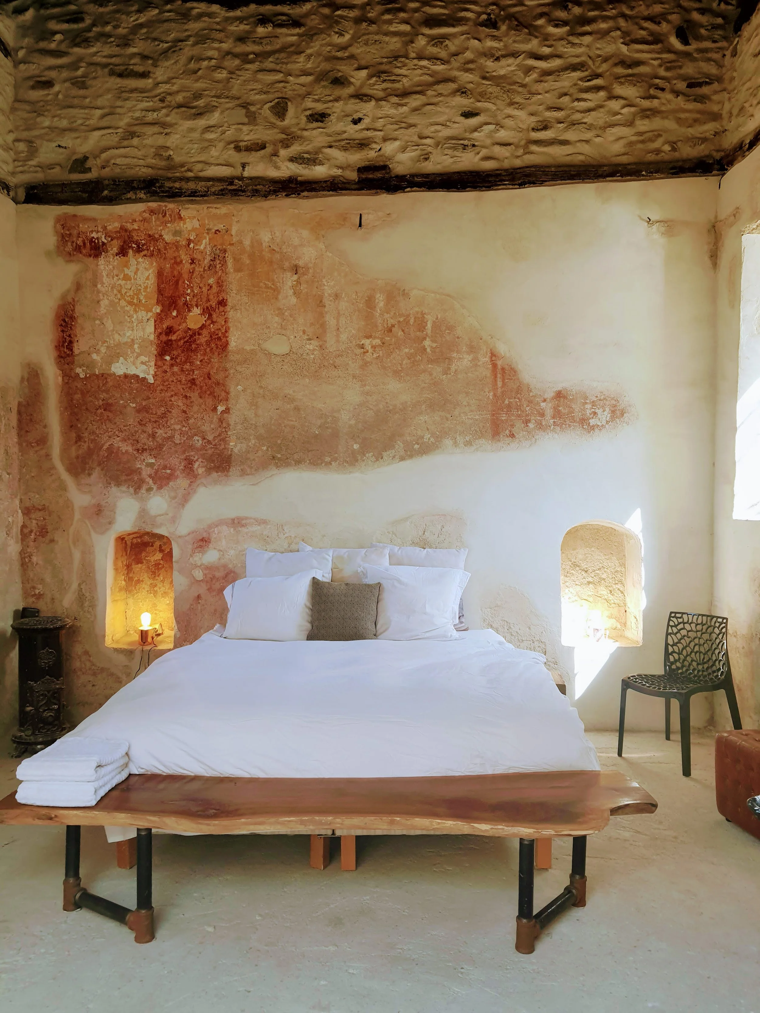A rustic bedroom with a bed featuring white linens and a small gray pillow, a wooden bench at the foot of the bed, and two small window alcoves with lighting. The wall behind the bed has a weathered, textured, partially exposed brick and plaster surface, and there is a black chair with a floral pattern on the right side.