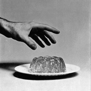 A hand reaching towards a plate with a gelatin mold in the center, black and white photograph.