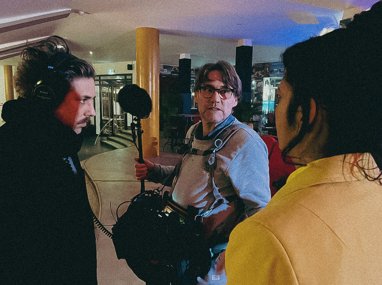 Drie mensen in een binnenruimte, twee daarvan lijken te filmen of fotograferen een man met een kabel en een audioapparaat. De man met het apparaat draagt een grijs shirt en heeft een rugzak. Een persoon draagt een gele jas en een ander persoon is gek