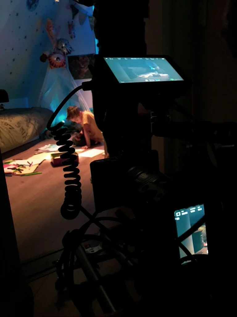A camera on a tripod filming a young girl in a bedroom with blue walls decorated with star patterns, sitting on the floor near a bed with a pillow and plush toys.