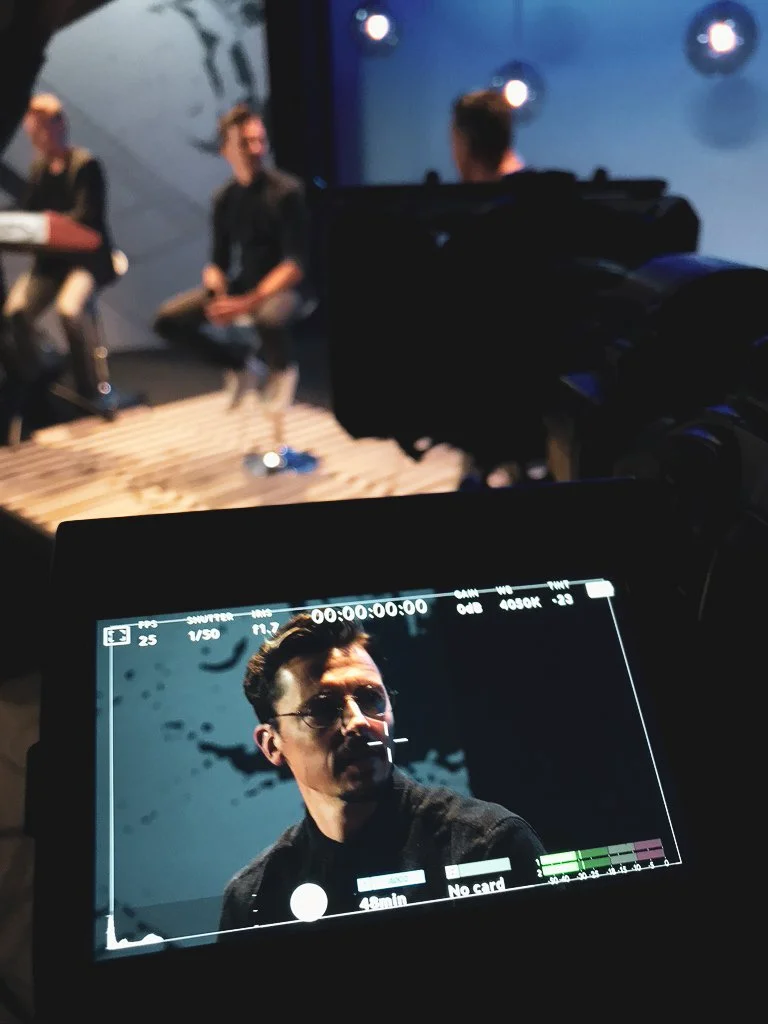 A person wearing glasses and a dark shirt is being filmed on a stage with a camera. Three other people are sitting on the stage, and the background is dark with some illuminated spots.