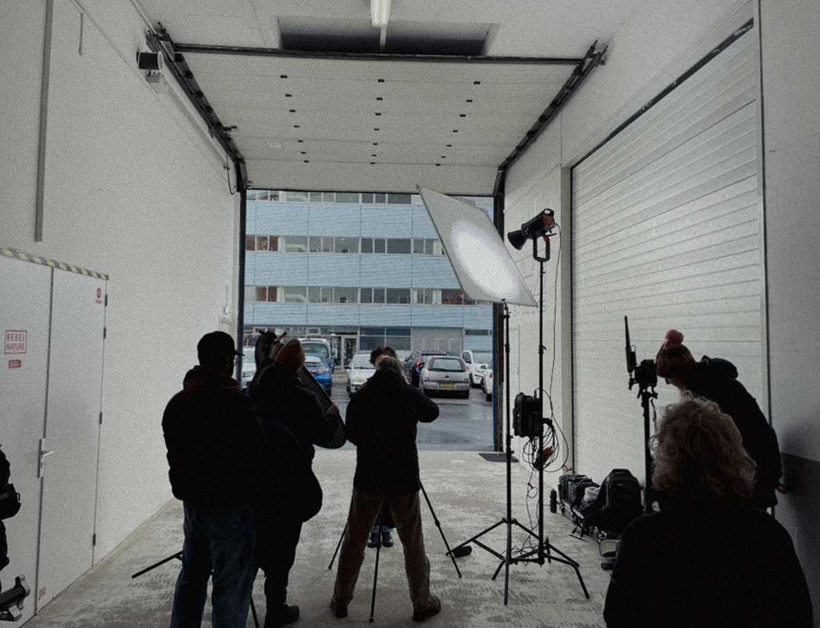 Binnen in een garage of studio met film- of fotoshoot-opstellingen, inclusief studioverlichting en apparatuur, met mensen die klaarstaan voor opname.