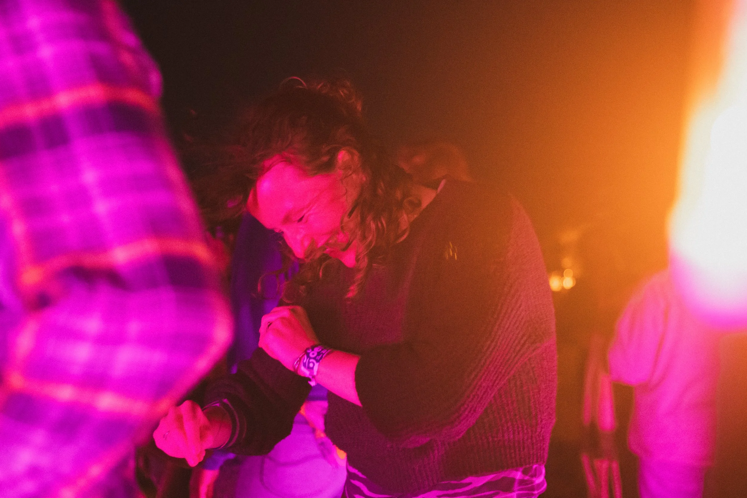 A woman with curly hair dancing or moving to music in a dimly lit environment with pink and orange lighting. Other people are partially visible around her.