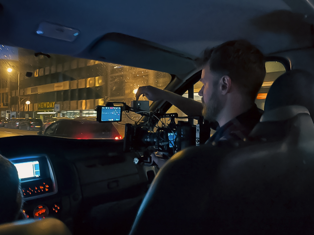 Binnenkant van een voertuig met iemand die een camera of filmingapparatuur bedient, nachtelijke stadsomgeving te zien door de ramen.