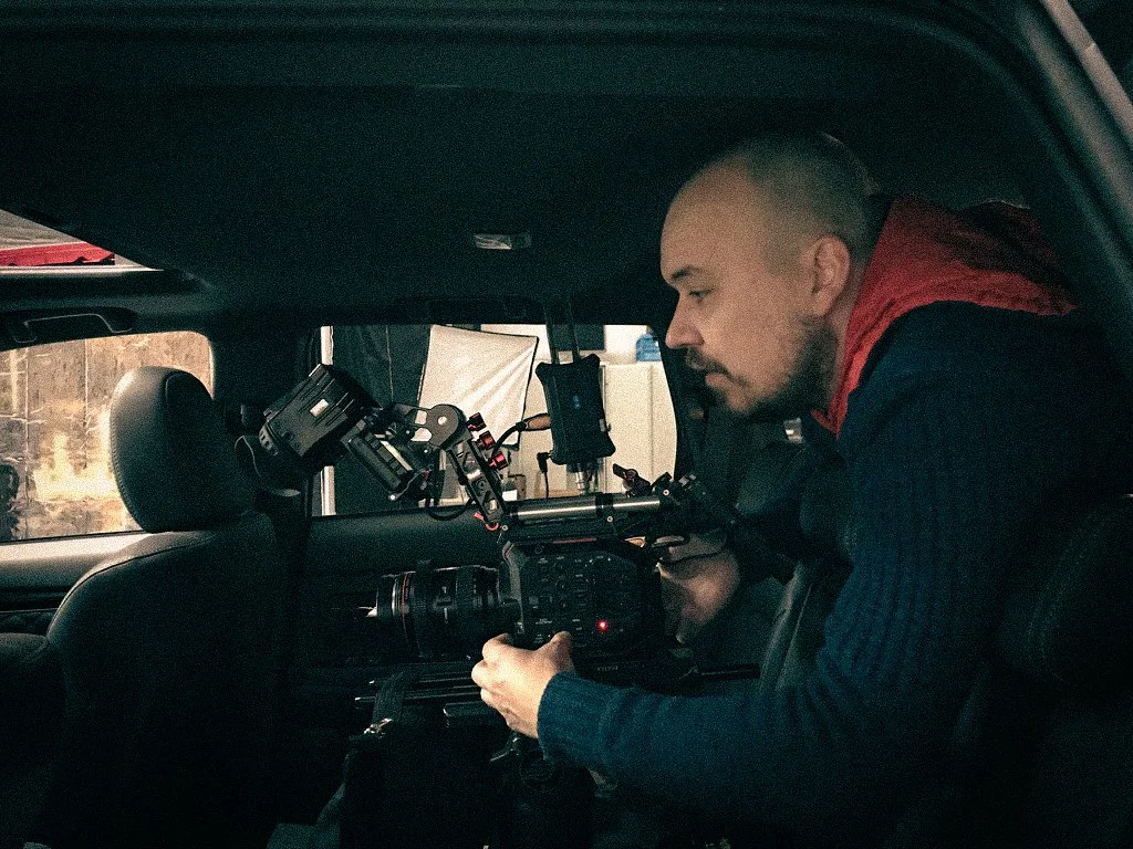A man with a beard and short hair is wearing a red and navy jacket, sitting inside a vehicle, working with professional camera equipment.