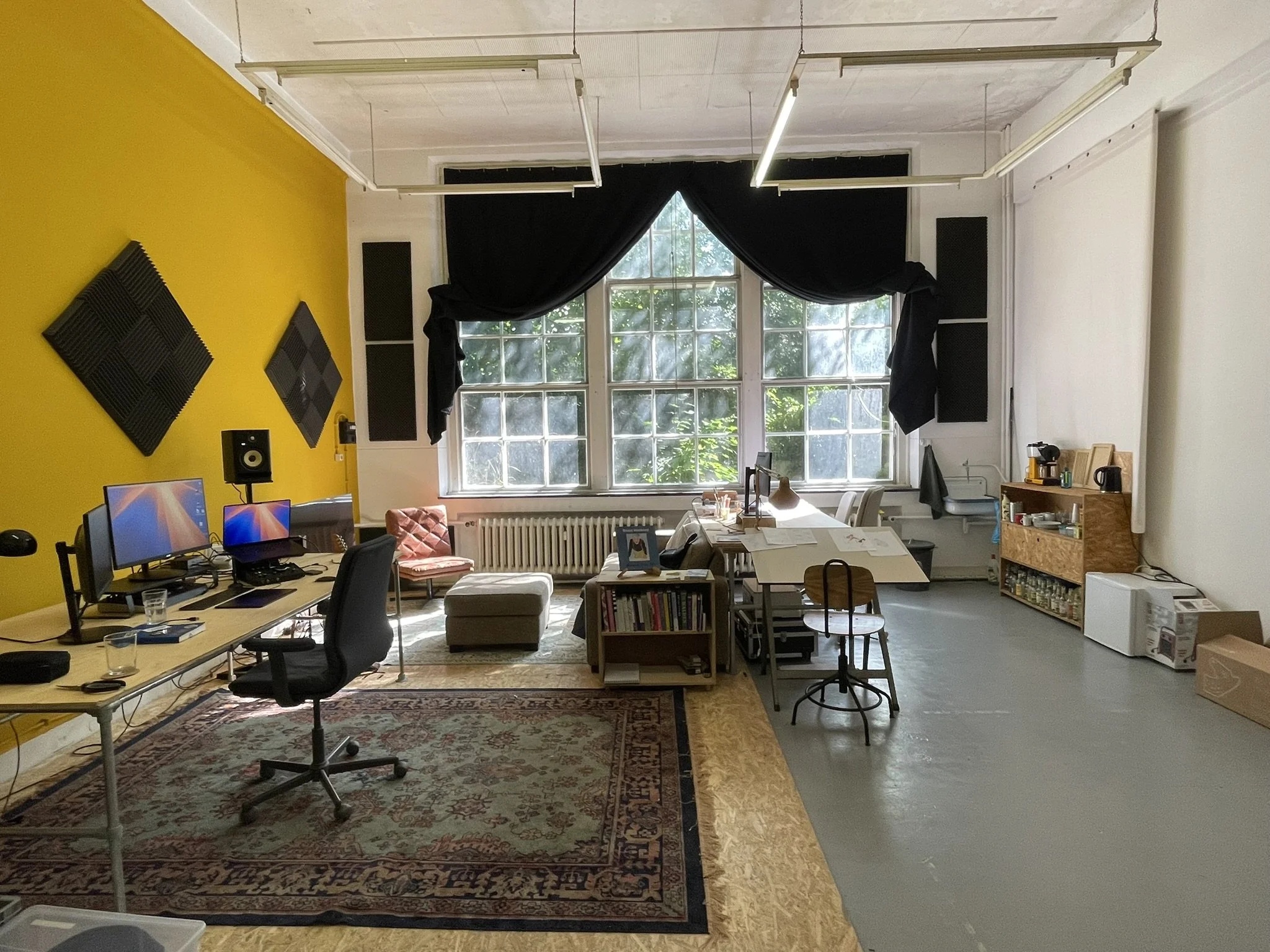 A spacious room with white and yellow walls, large window with black curtains, multiple computer monitors on a desk, soundproofing foam panels, a cozy corner with a pink cushioned chair and ottoman, and various office and kitchen items.