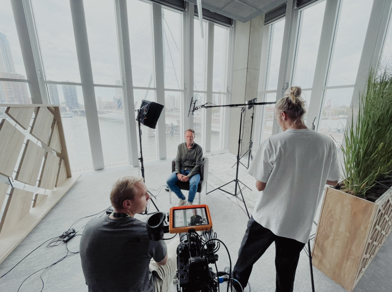 Een interview of opname sessie met een man die voor grote ramen zit met uitzicht op een rivier en stadsgezicht, terwijl twee mensen achter de camera de opname maken.