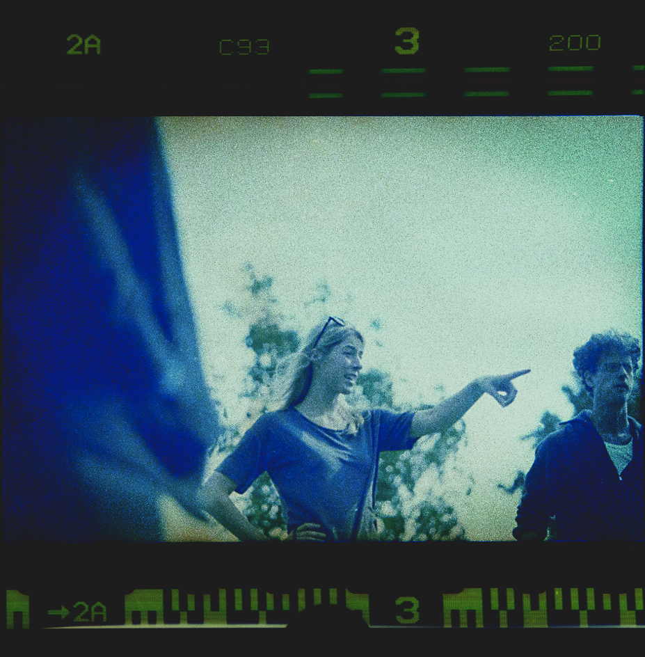 Een vrouw wijst naar rechts, terwijl ze met twee andere mensen buiten staat, onder een heldere hemel met bomen op de achtergrond.