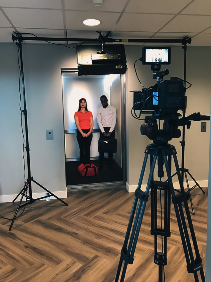 Twee mensen wachten in een lift, gefilmd met een camera en belichtingsapparatuur in een studio-achtige omgeving.
