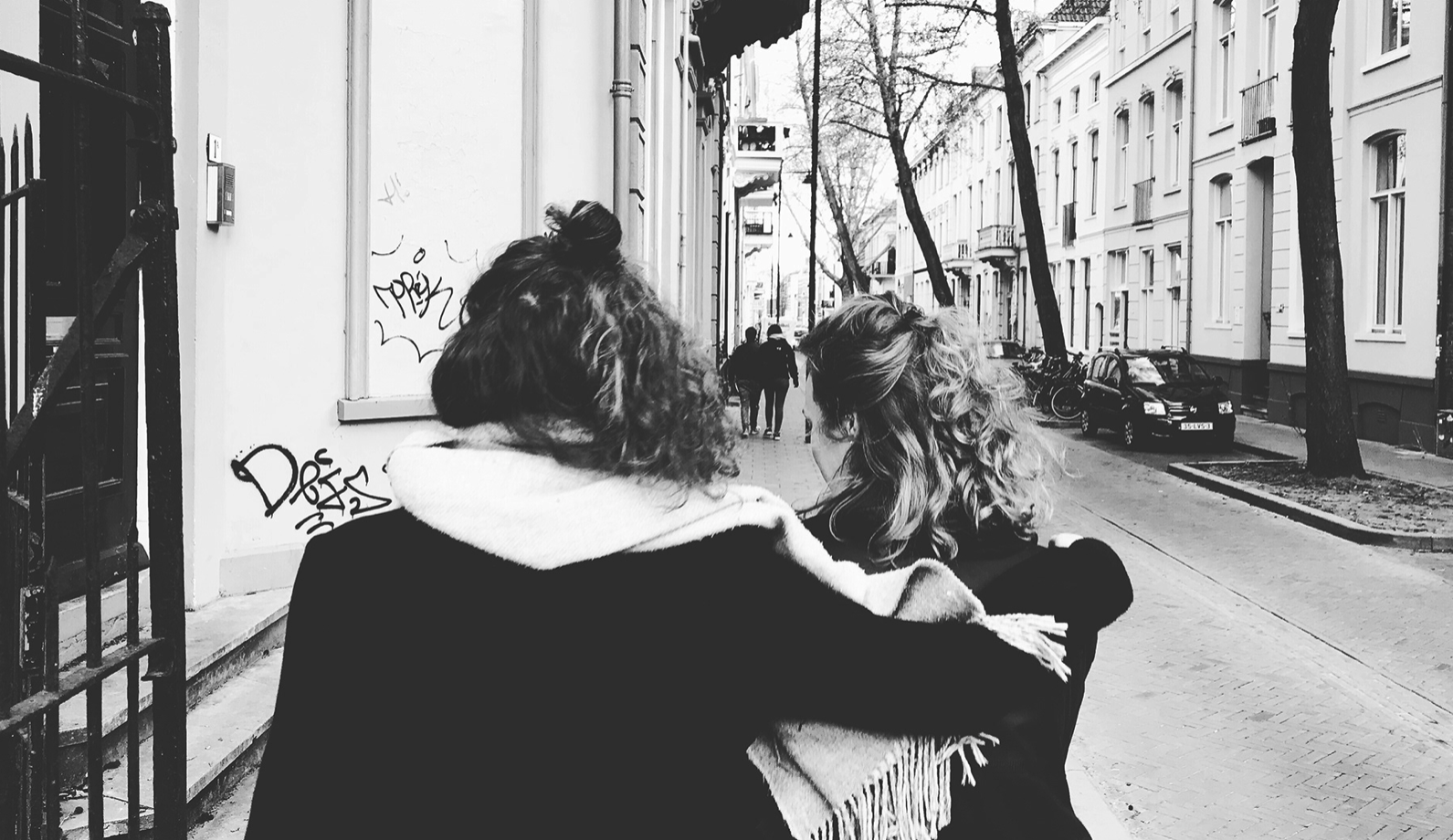 Twee vrouwen lopen hand in hand op een stadswandeling, de straat is omringd door gebouwen en bomen, in zwart-wit.