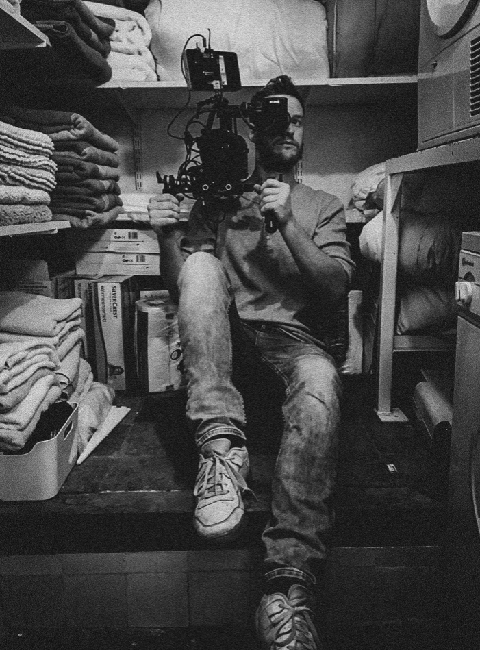 Man zit op de vloer in een kast met stapels handdoeken en huishoudelijke spullen, neemt een foto met een professionele camera.