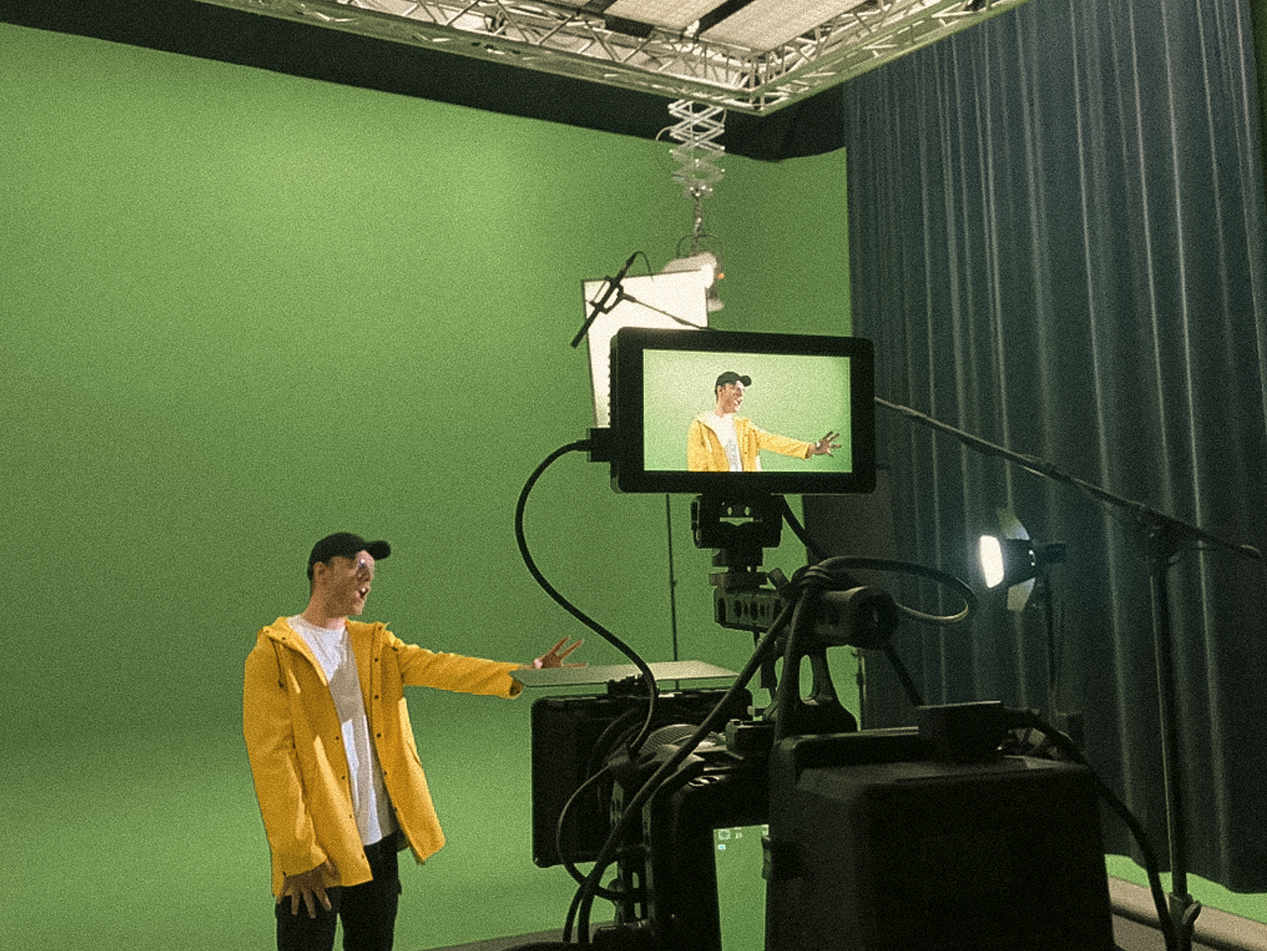 Een man in een gele jas poseert voor een groene scherm in een filmstudio, terwijl hij uitbeeldt met een handgebaar. Er is cameraapparatuur en een monitor die zijn afbeelding weergeeft.