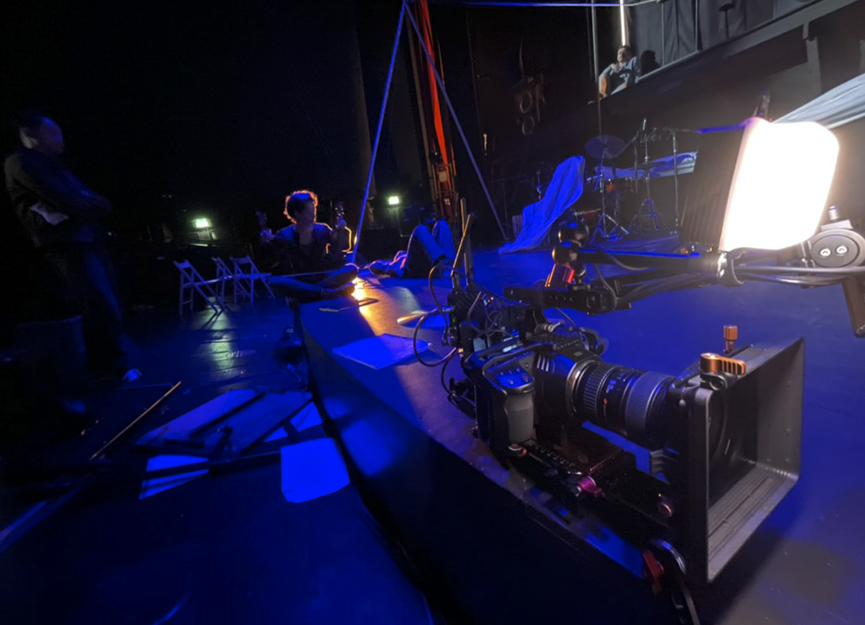 Camera op een filmset met mensen en equipment, donker met blauwe verlichting die het toneel verlicht.