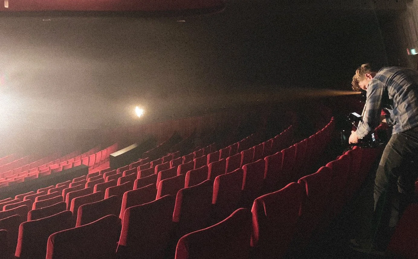 A person operating a camera backstage in an empty movie theater with red seats and a bright light in the background.