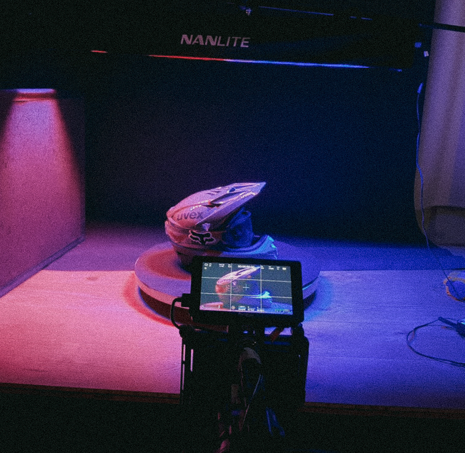 Een racehelm op een ronde platform, met een camera in de voorgrond en neonlichten op de achtergrond met het woord 'NANLITE'.