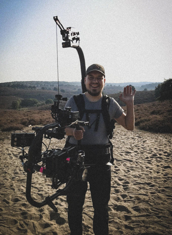 Man met camerastabilizer in een rivieromgeving, zwaaiend en lachend naar de camera.